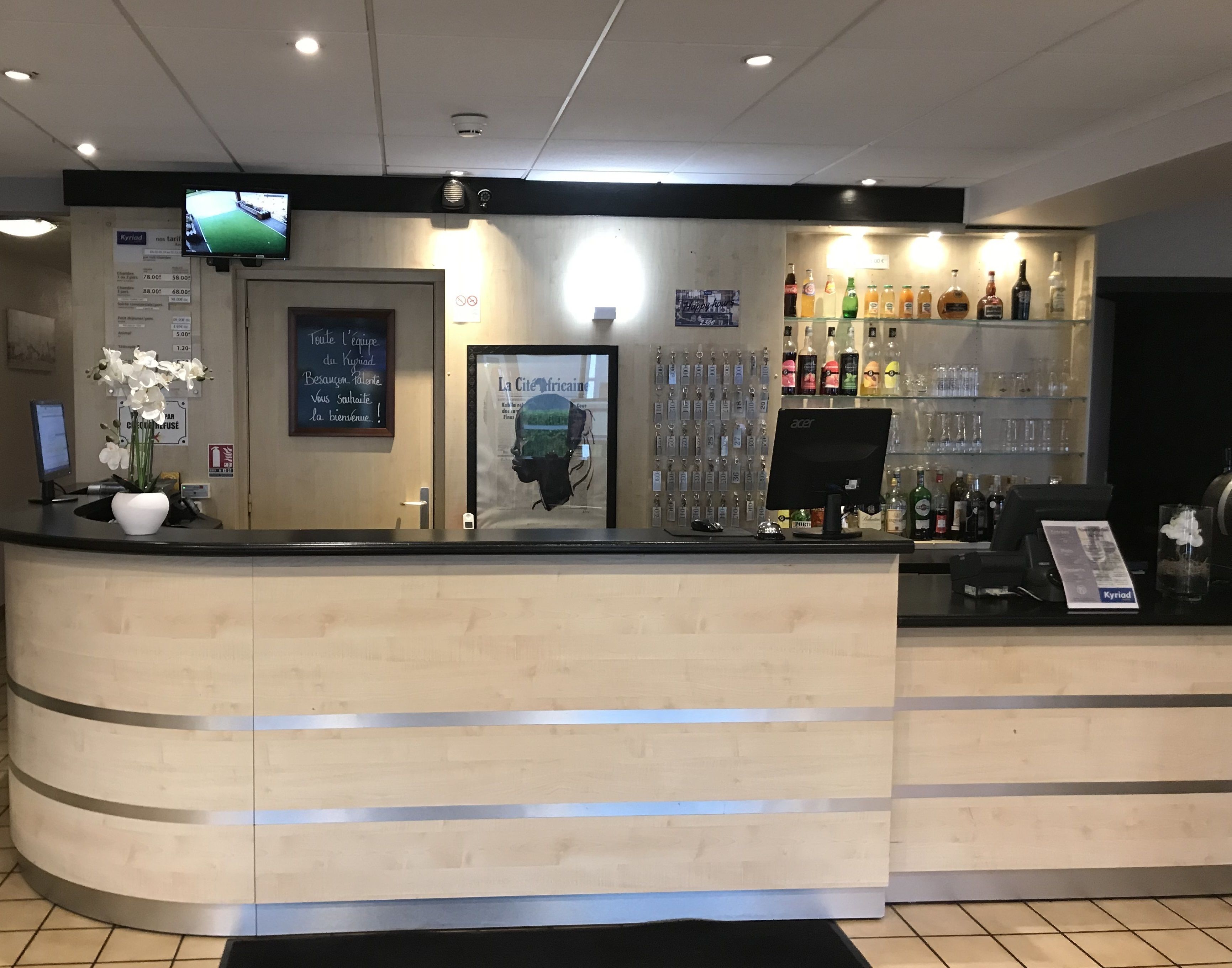 A hotel reception area with a curved counter, a mounted television, a framed picture, a selection of bottled beverages, and a computer monitor.