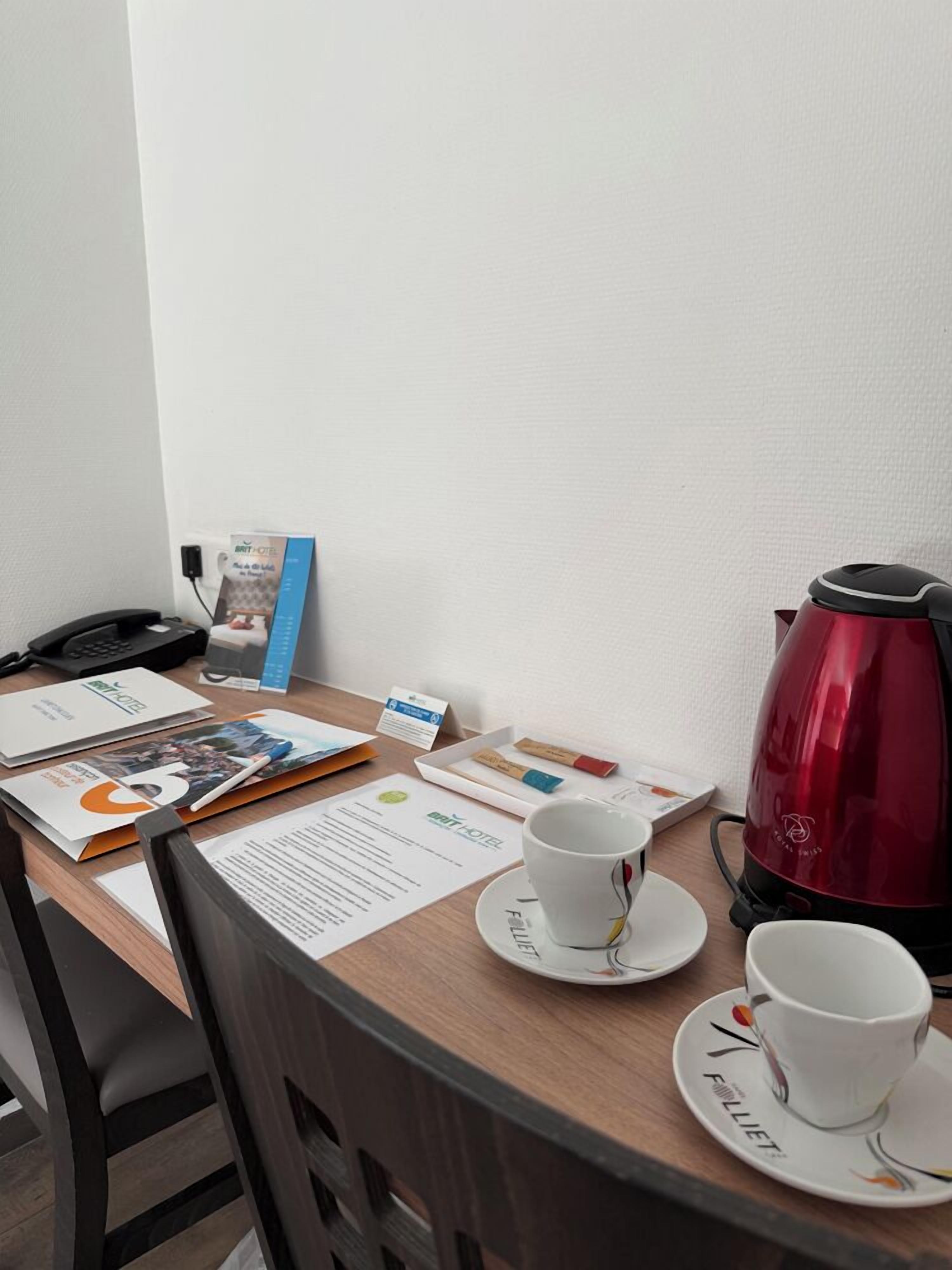 A hotel room with a wooden desk, a red kettle, two cups, a phone, and various papers and a brochure.