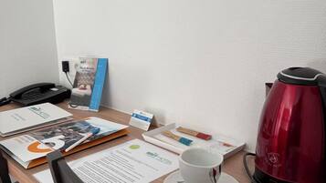 A hotel room with a wooden desk, a red kettle, two cups, a phone, and various papers and a brochure.