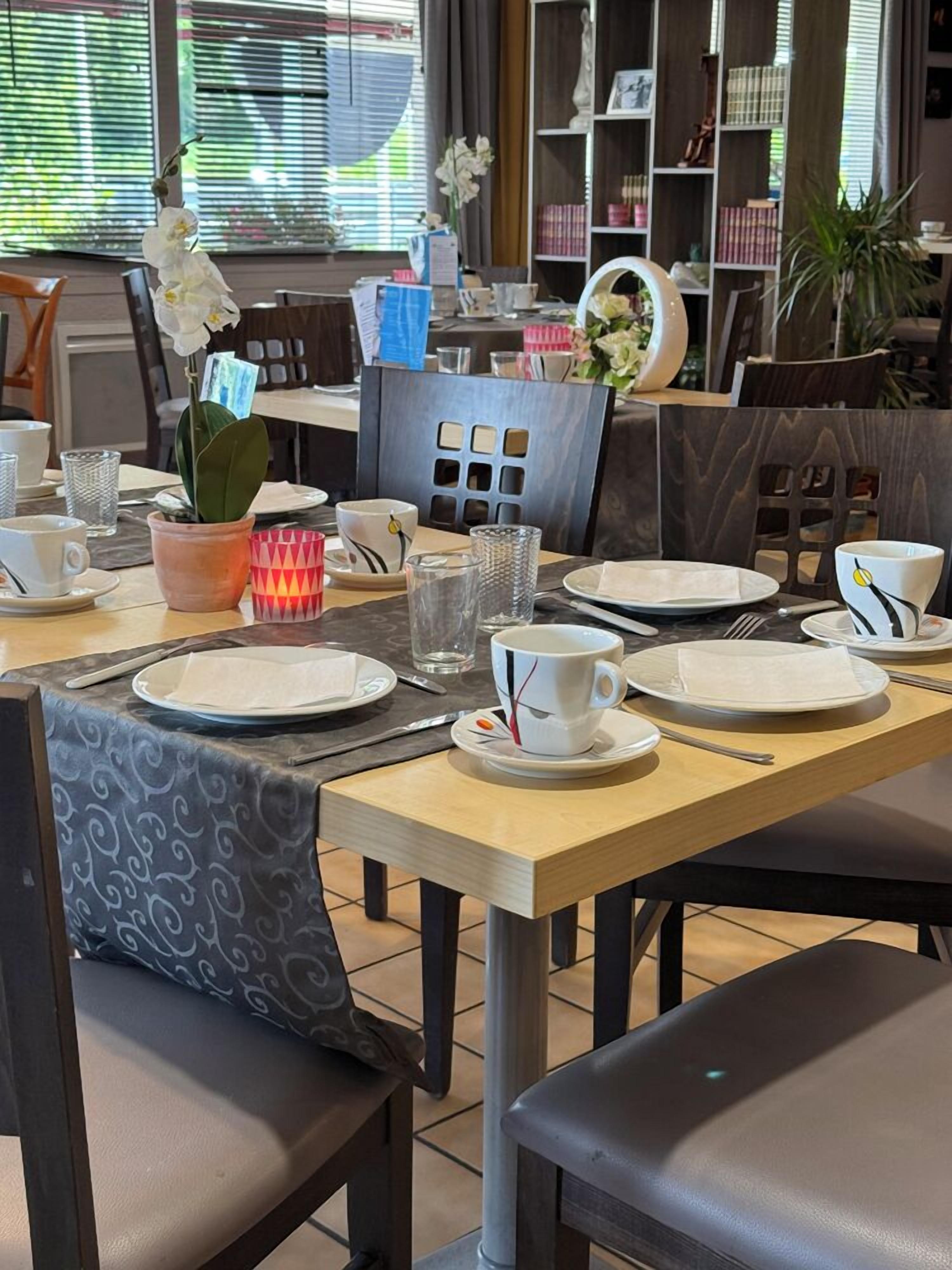 A dining area with a table set for two, chairs, a potted plant, and a bookshelf in the background.