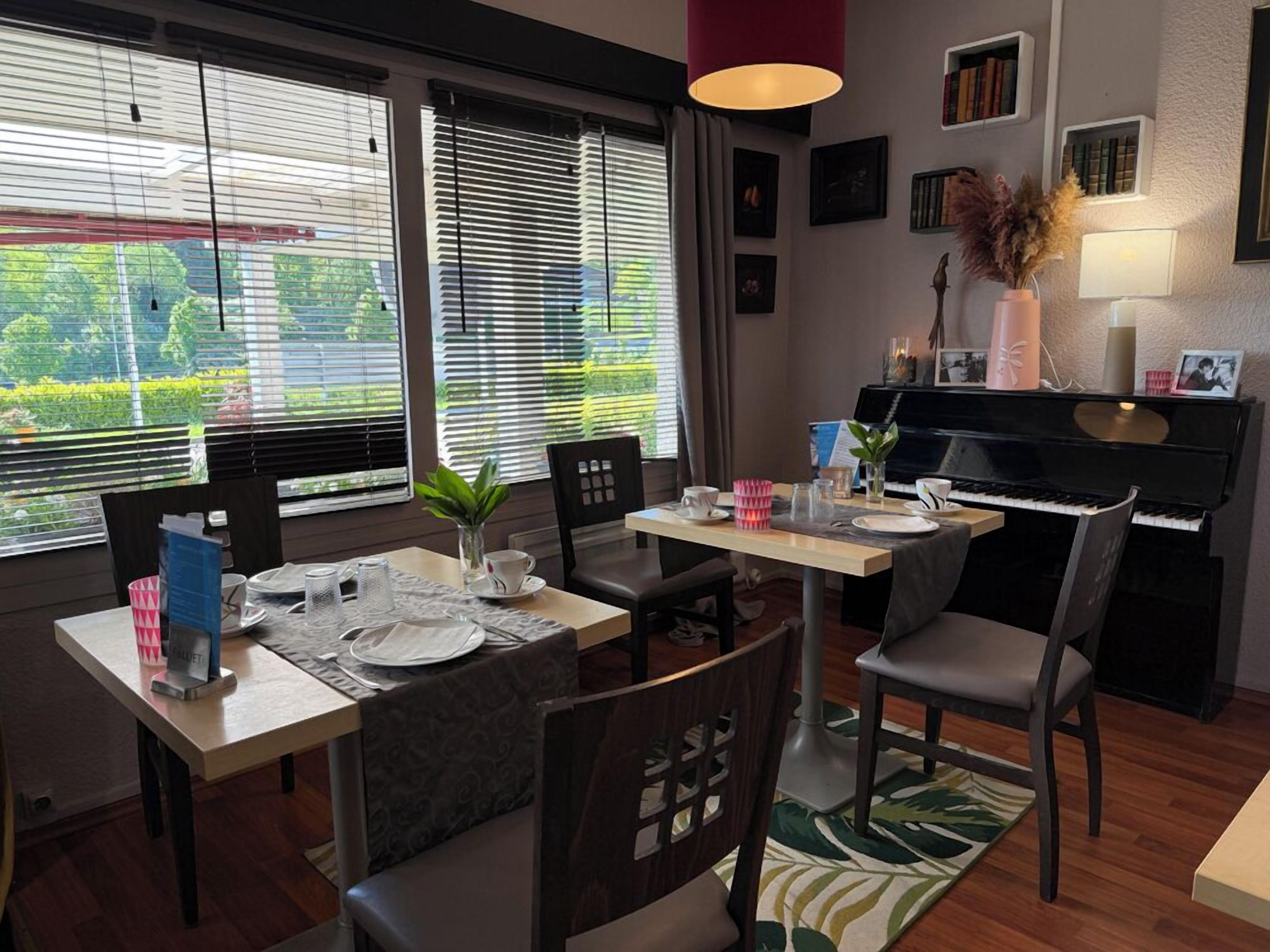 A dining area with a piano, dining table, chairs, and a view through large windows.