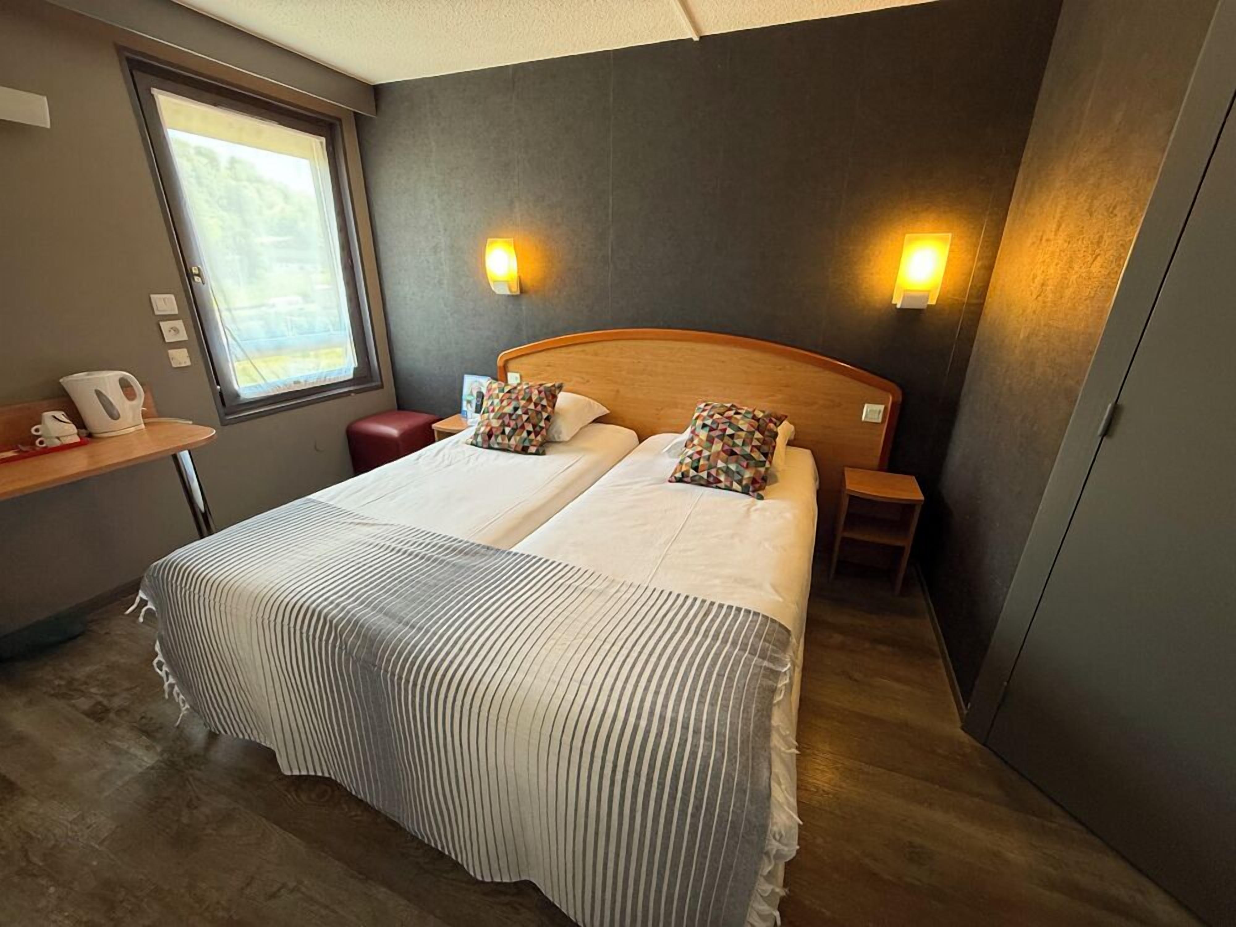 A double bed with striped bedding, two bedside tables, a nightstand, a window with a view, and a wall-mounted light fixture.