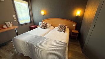 A double bed with striped bedding, two bedside tables, a nightstand, a window with a view, and a wall-mounted light fixture.