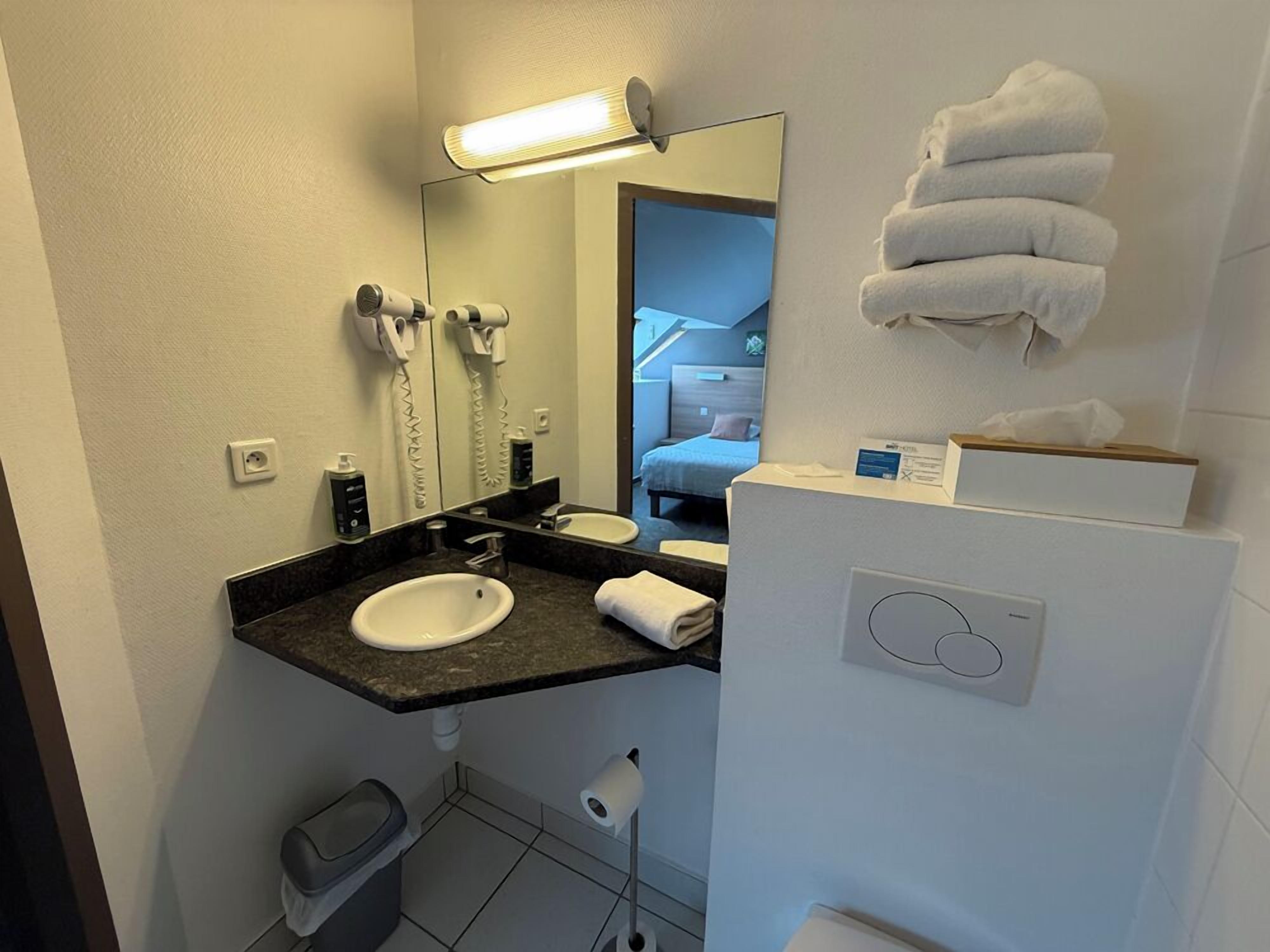 Un baño de hotel pequeño con un lavamanos, dos secadores de pelo, un espejo, un toallero y un inodoro.