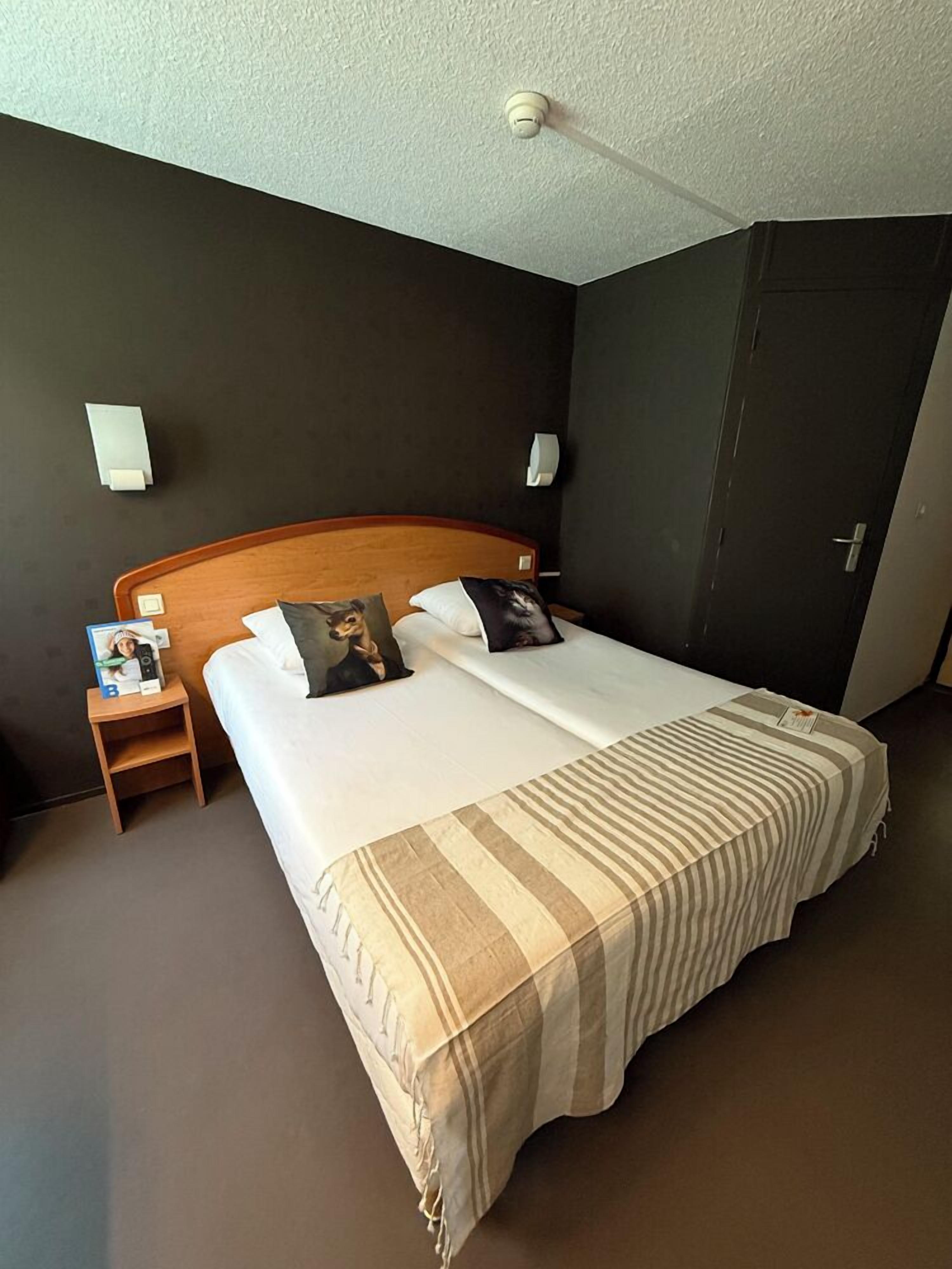 Habitación de hotel con cama doble, dos mesitas de noche, un aparador, una lámpara fijada a la pared, una puerta y un detector de humo instalado en el techo.