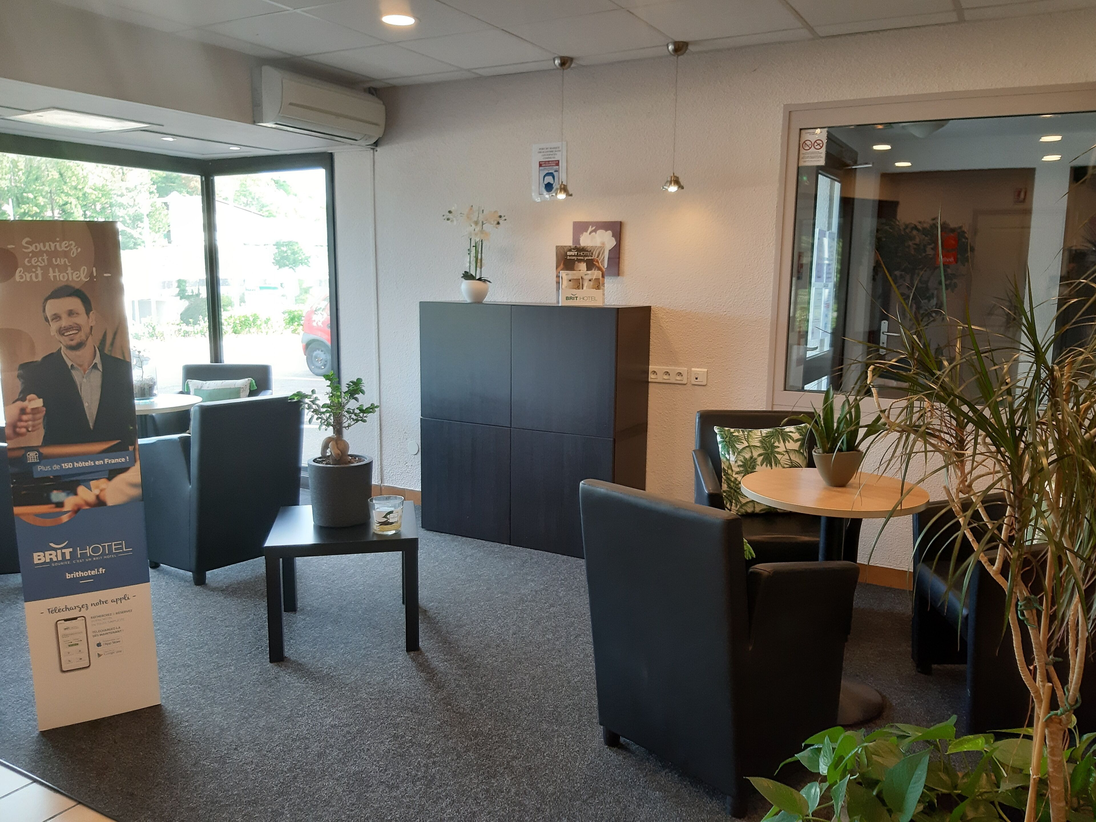 Et hotelreceptionsområde med en glasdør, en reception, en potteplante, et lille bord med et glas og en plakat der reklamerer for hotellet.