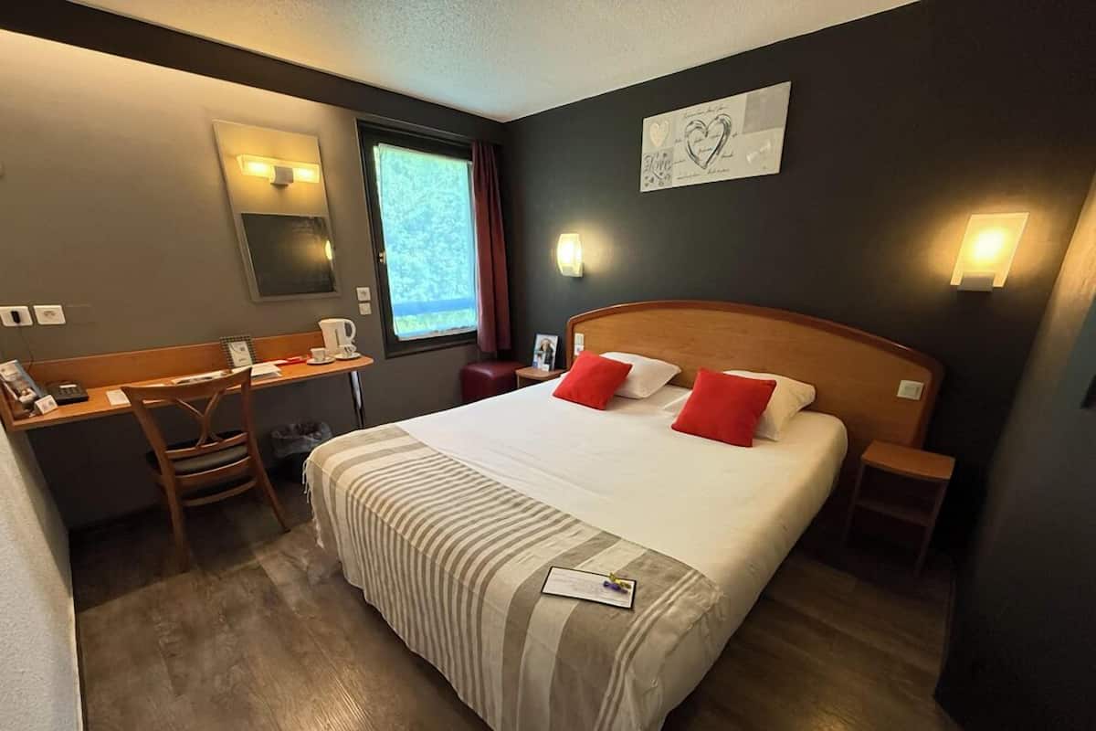 Habitación de hotel con una cama grande, un escritorio con silla, una mesita de noche, una ventana con cortinas e iluminación en la pared.