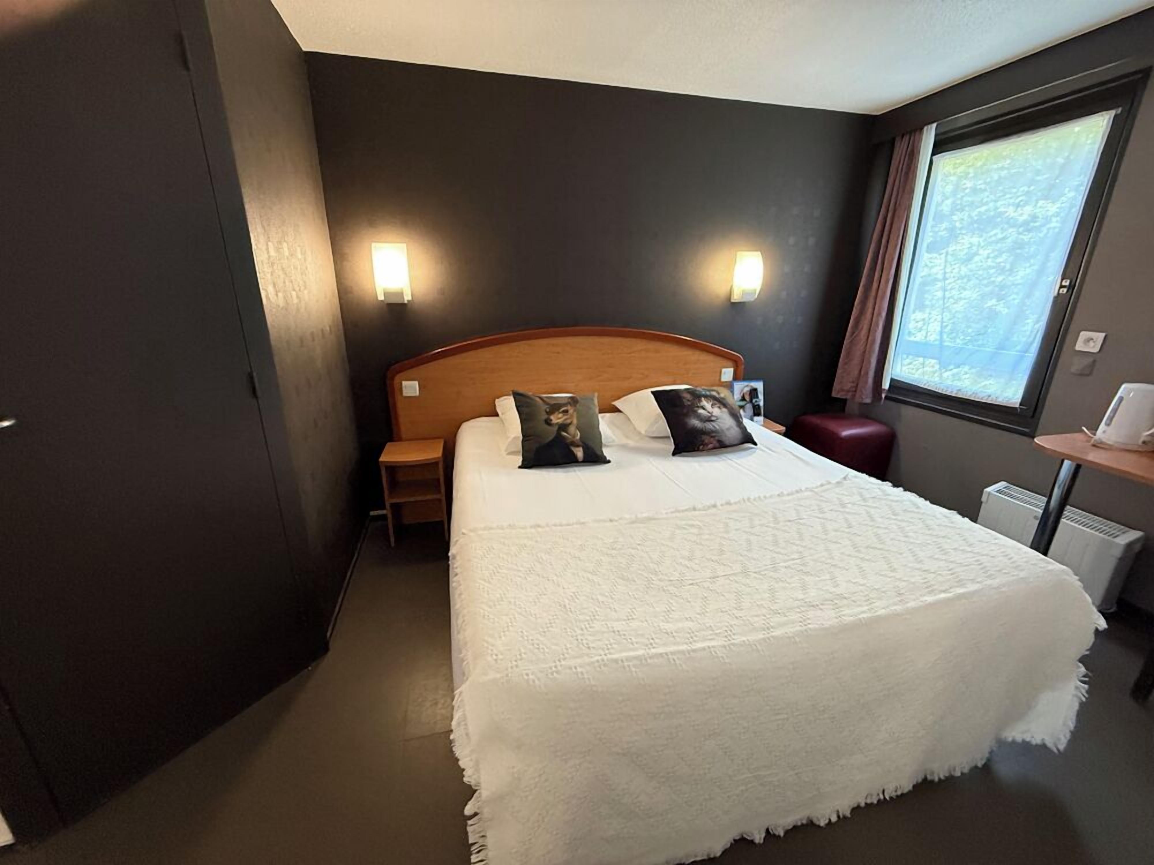 A hotel room with a large bed, two bedside lamps, a nightstand, a window with a view, and a red bench.