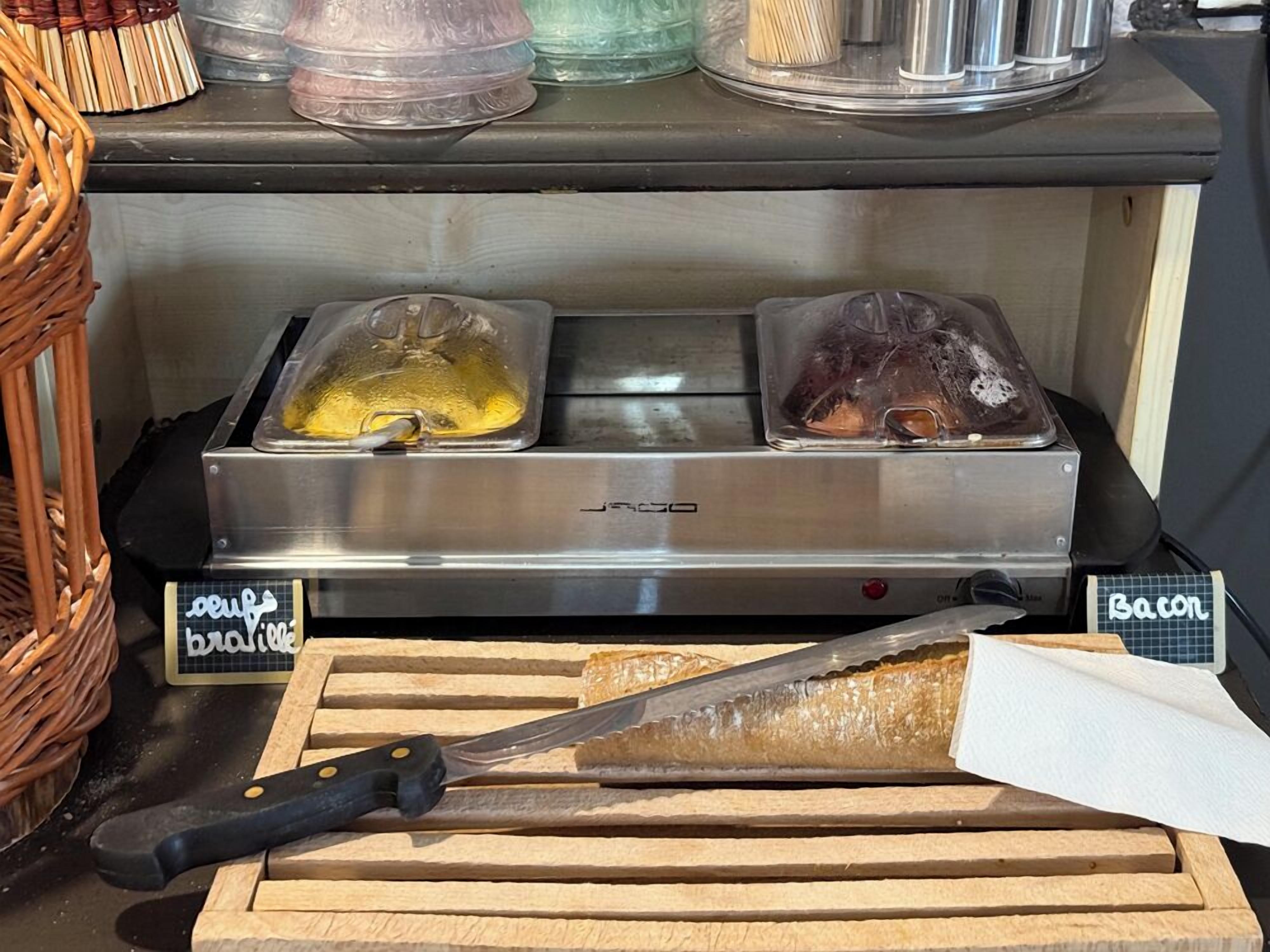 Ein Küchenbereich mit einem Herd der Marke J.R. Jones, zwei abgedeckten Speiserausgabesystemen, einem Schneidebrett mit Messer und einem Schild, das 'Bacon' und 'Brie' ausweist.