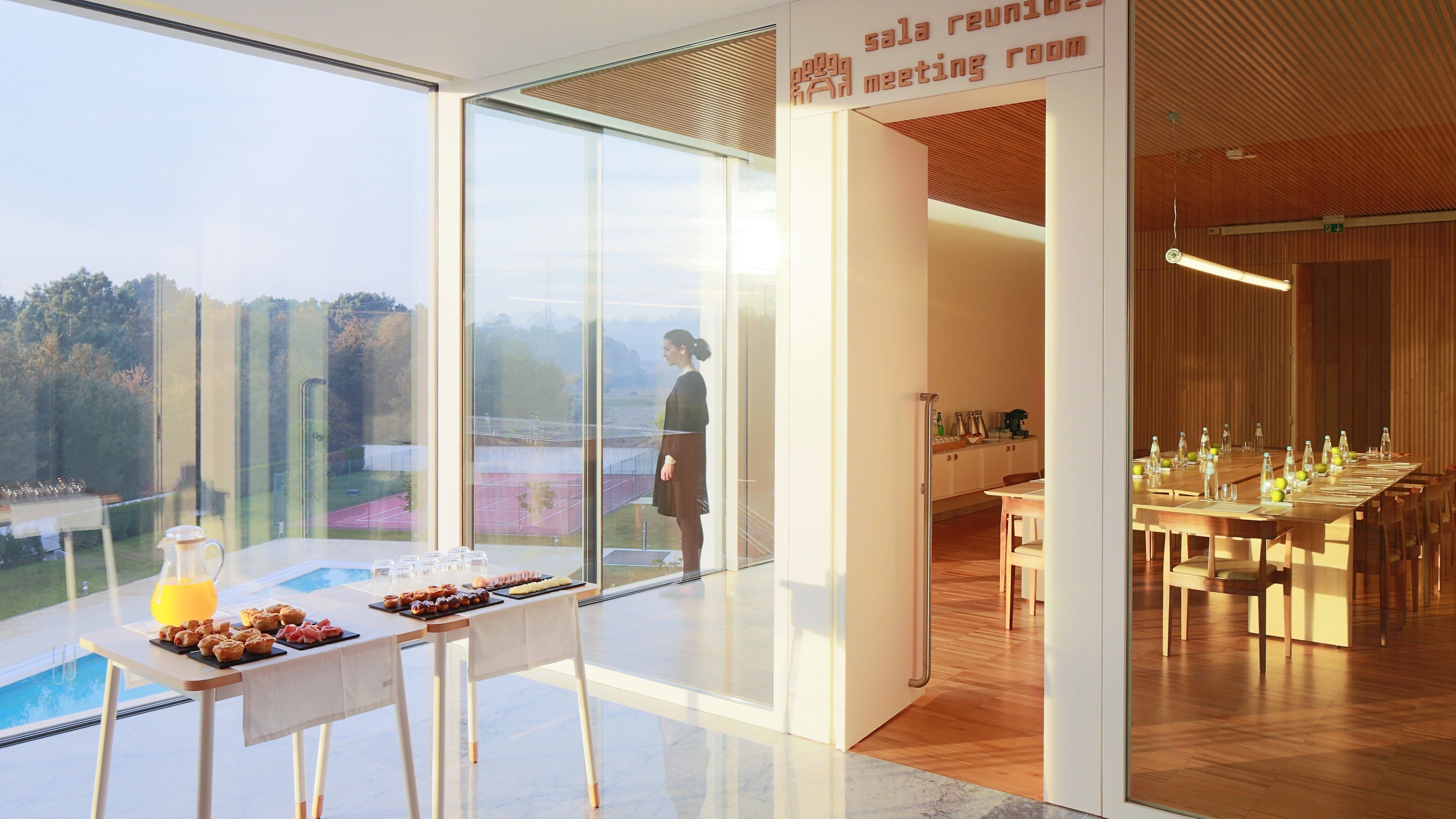 Una habitación de hotel moderna con un amplio muro de vidrio, un comedor con una mesa larga dispuesta para una comida, y un cartel que indica una sala de reuniones.