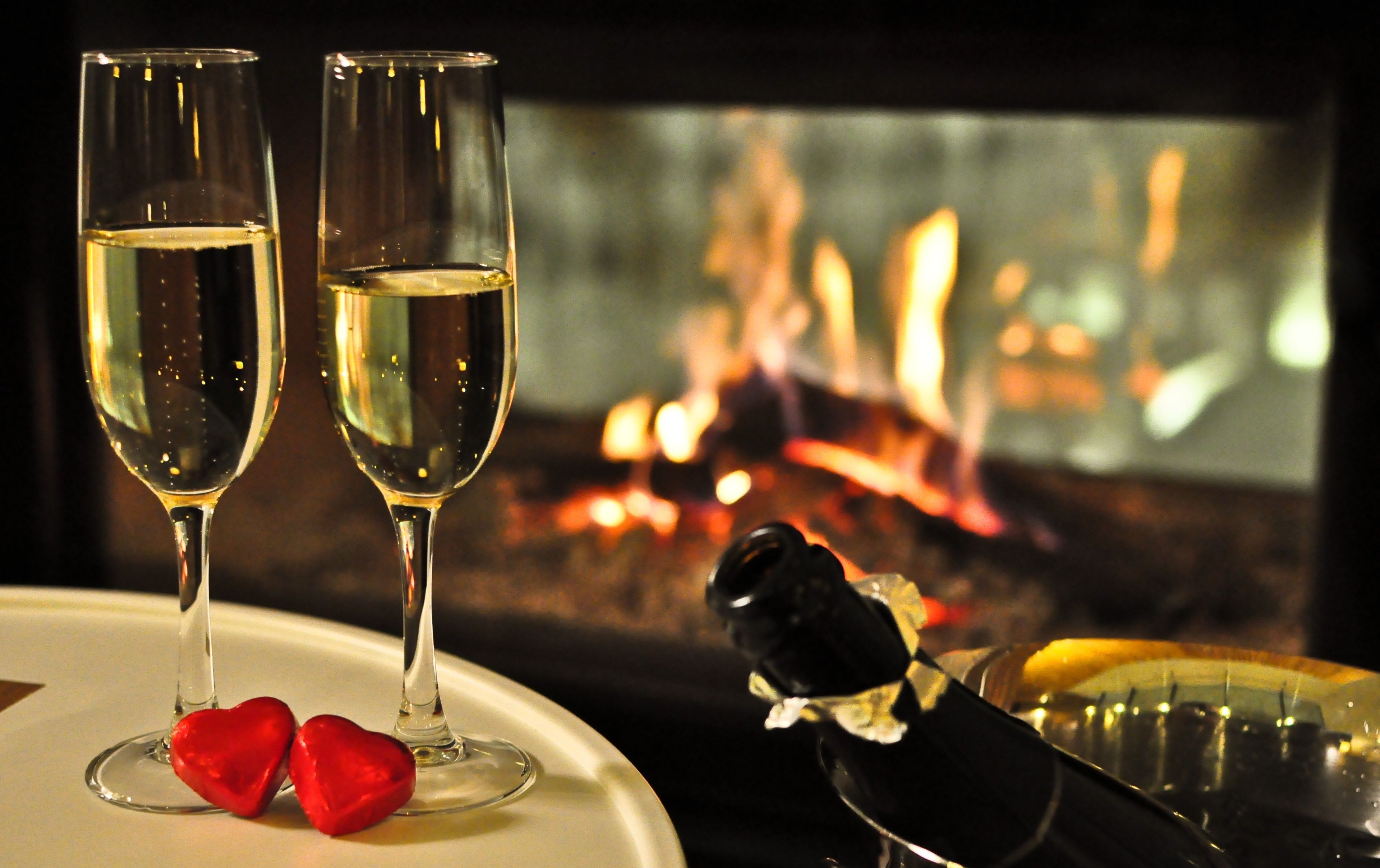 Two glasses of white wine on a table with red heart-shaped decorations, a bottle of champagne, and a fireplace in the background.