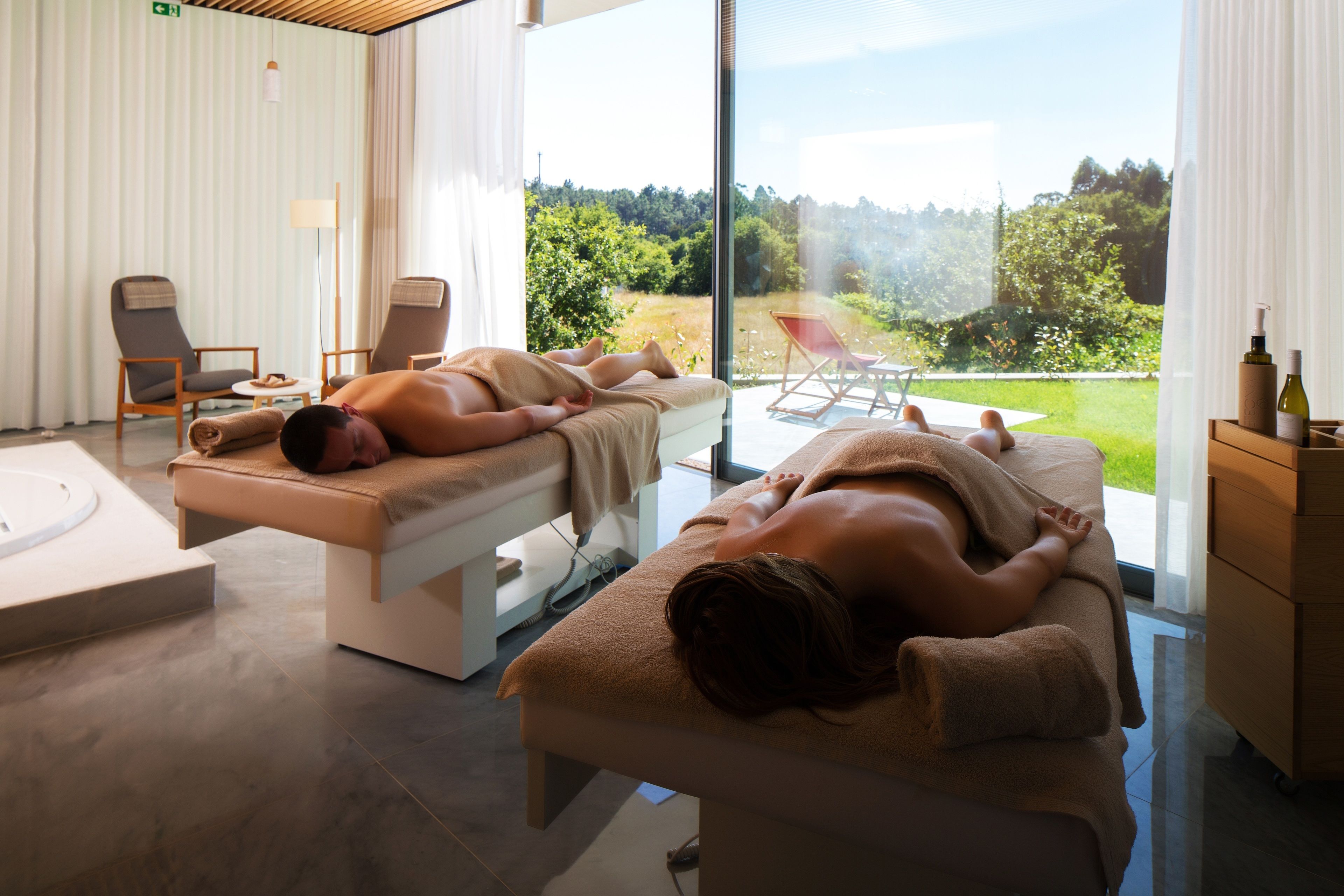 A spa room with two massage tables, a small table with a bottle and a bottle of lotion, and a view of a garden through large windows.