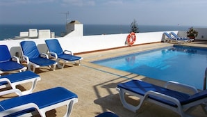 Zona junto a la piscina con sillas reclinables azules, un flotador salvavidas y vistas al mar.