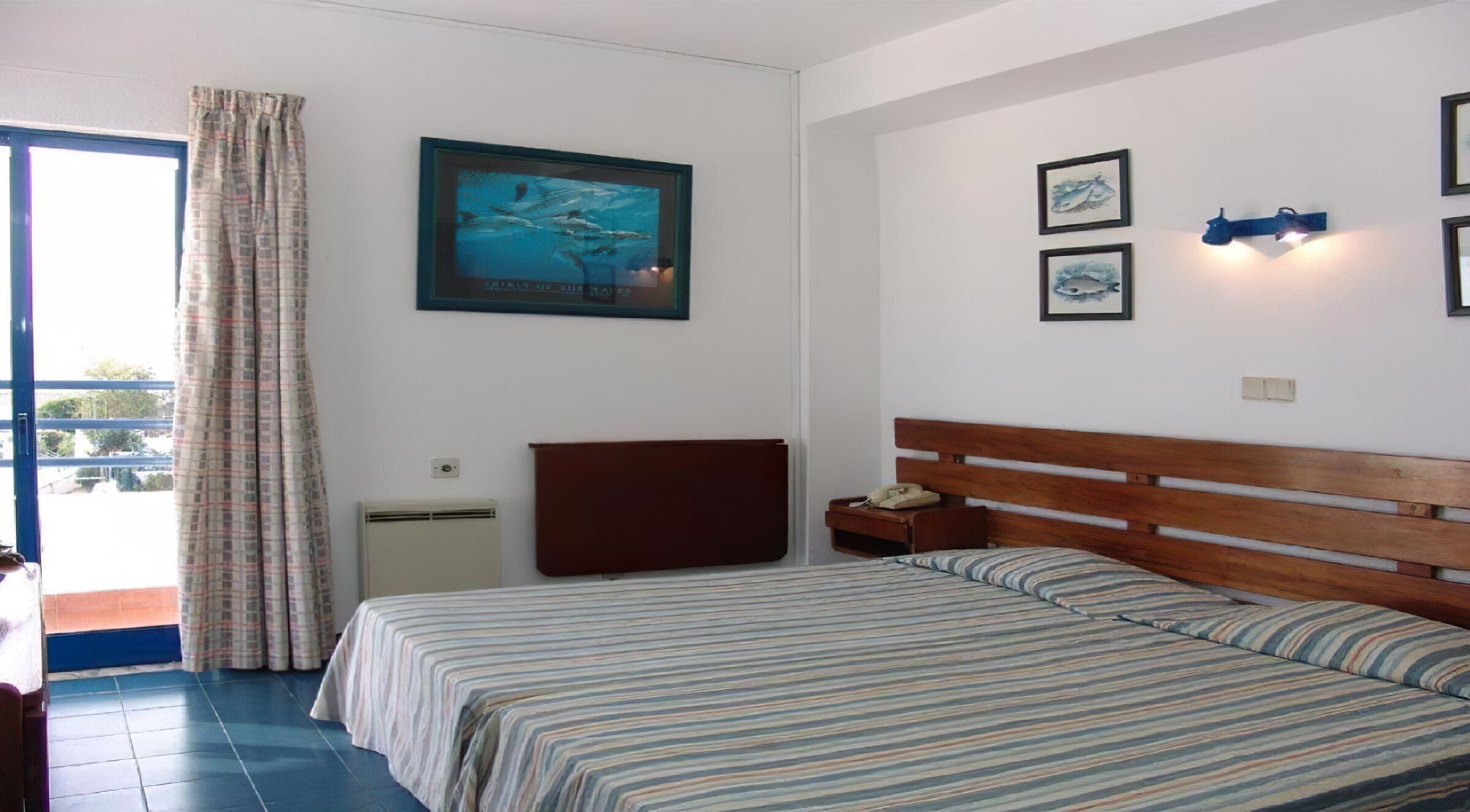 Une chambre à coucher comprenant un lit, une table de chevet, un tableau encadré, une lampe fixée au mur et une fenêtre avec des rideaux.
