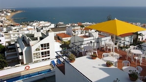 Una terraza en la azotea con una sombrilla amarilla, sillas blancas y plantas en macetas.