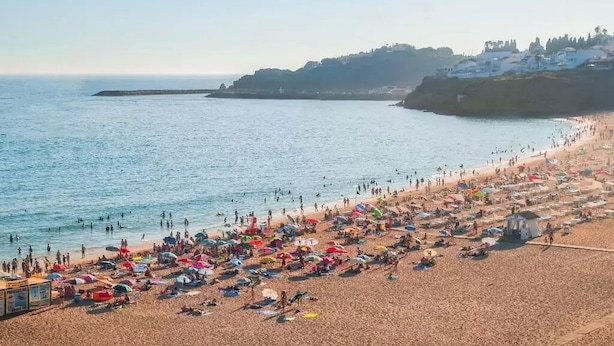 Una playa con gente tomando el sol y nadando, un cielo despejado y edificios en una colina al fondo.