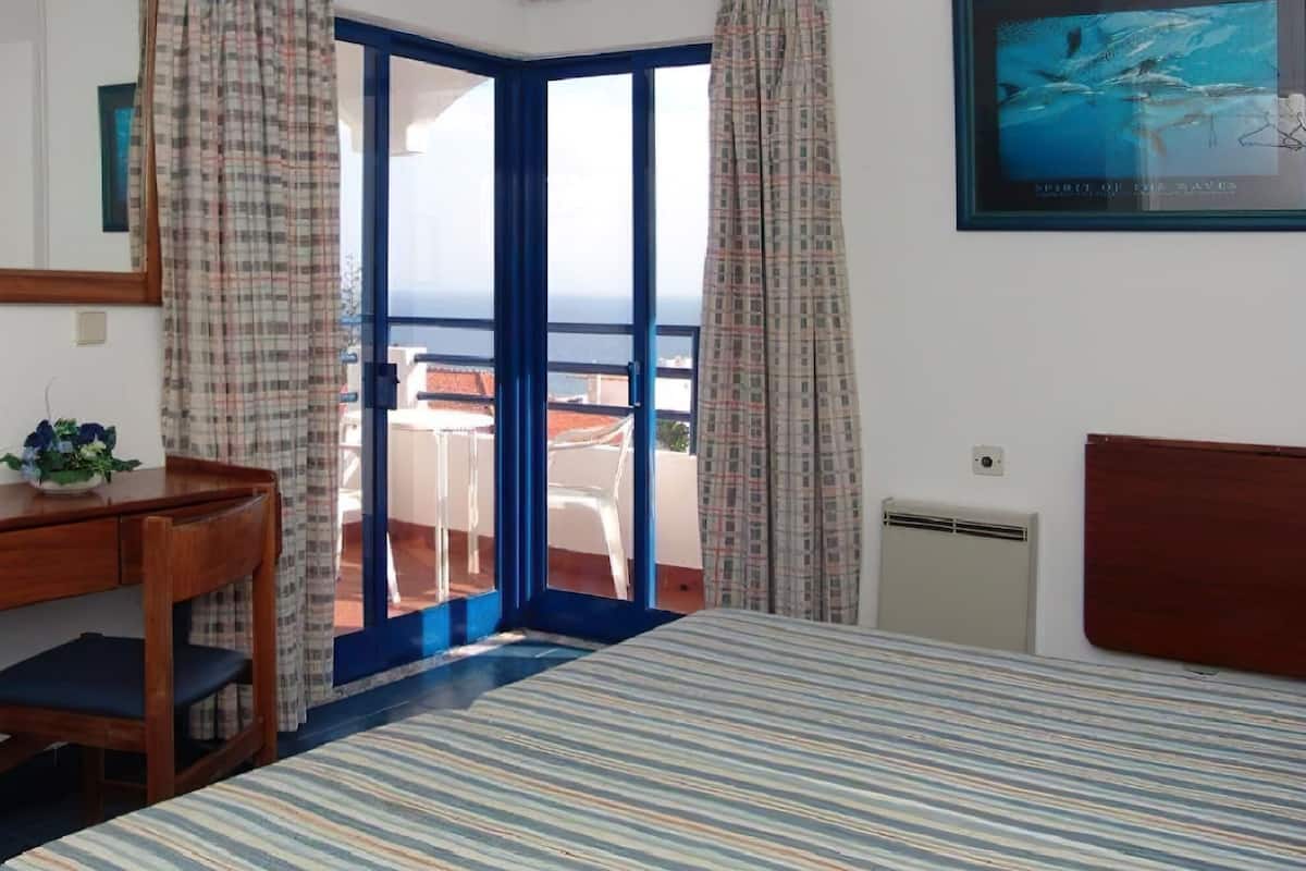 A hotel room with a bed, a desk, a chair, a window with curtains, and a framed picture on the wall.