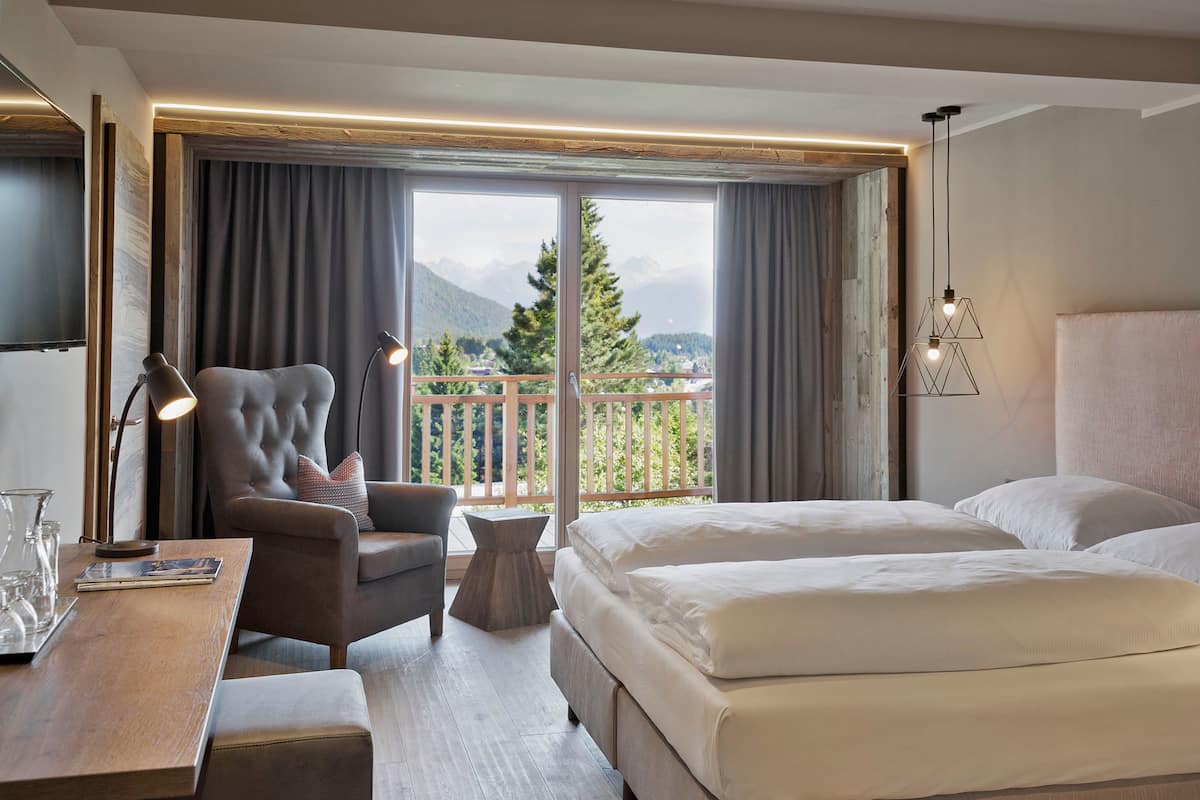 A hotel room with a large bed, a desk with a lamp, a chair, and a view of mountains through the window.