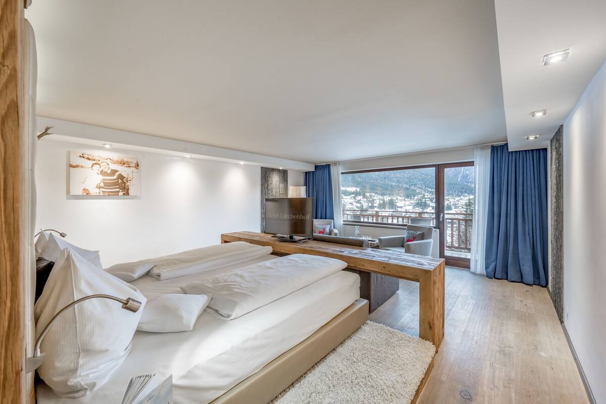 A modern hotel room with a large bed, a desk, a TV, and a balcony with a view of the mountains.