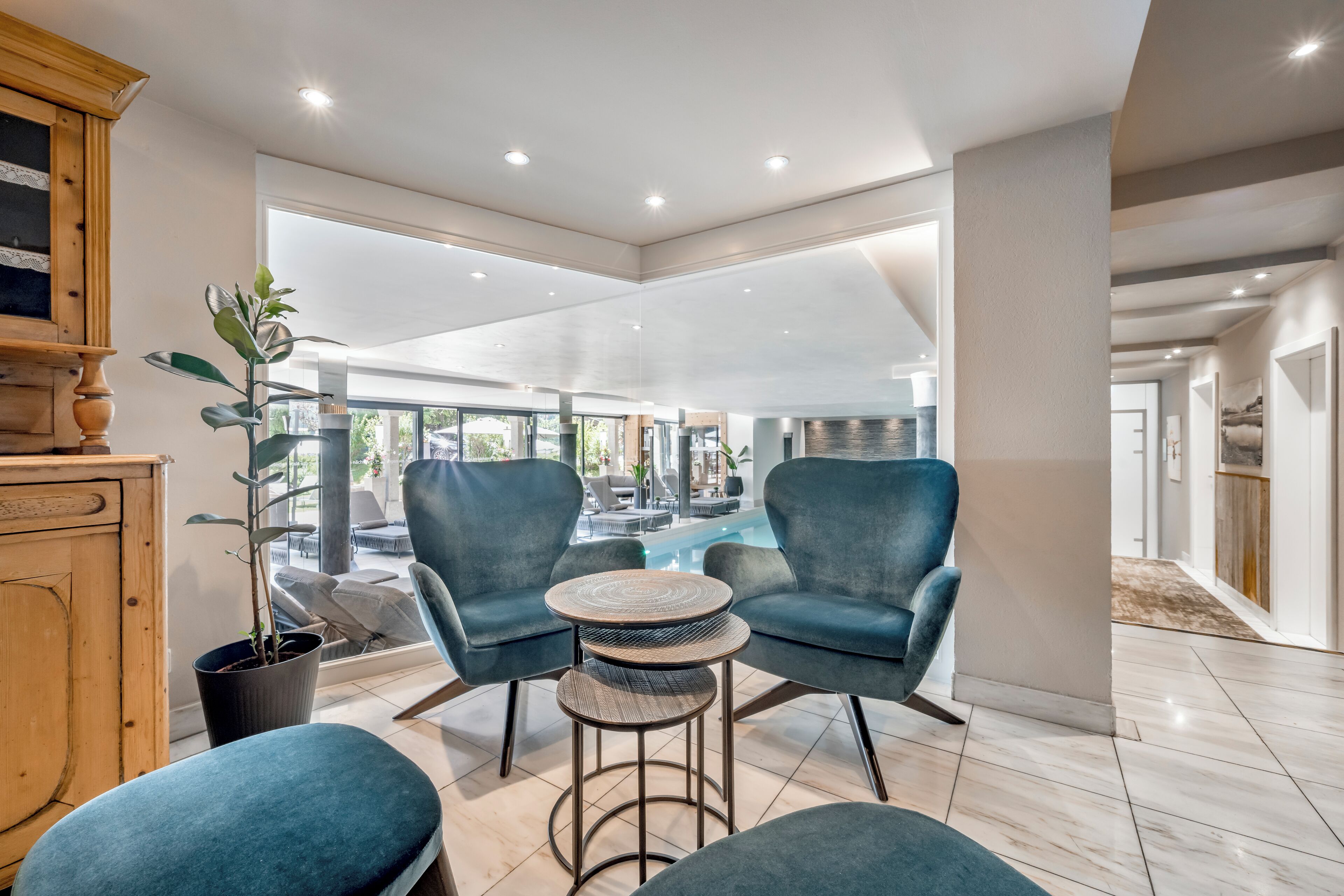 Un moderno salón con vista a la piscina, con dos sillones azules, una mesa redonda y una planta en maceta.