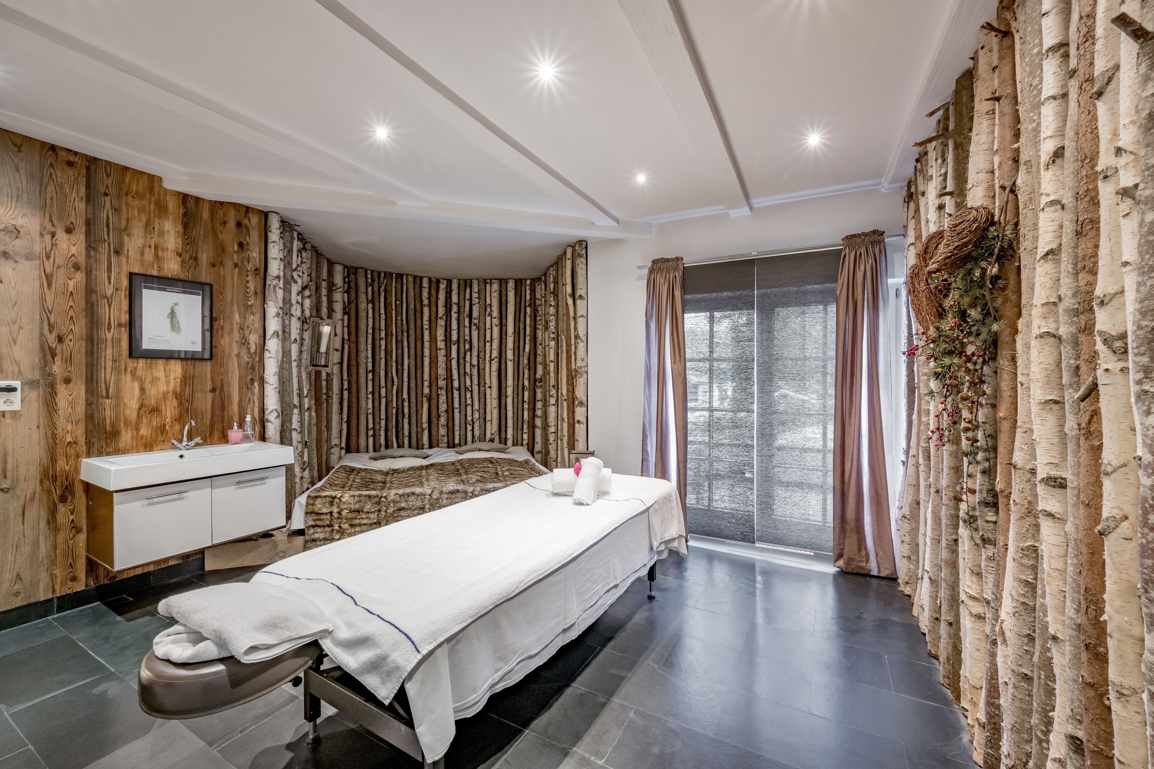 A modern spa room with a wooden wall, a large bed, a white sink, and a large window with curtains.