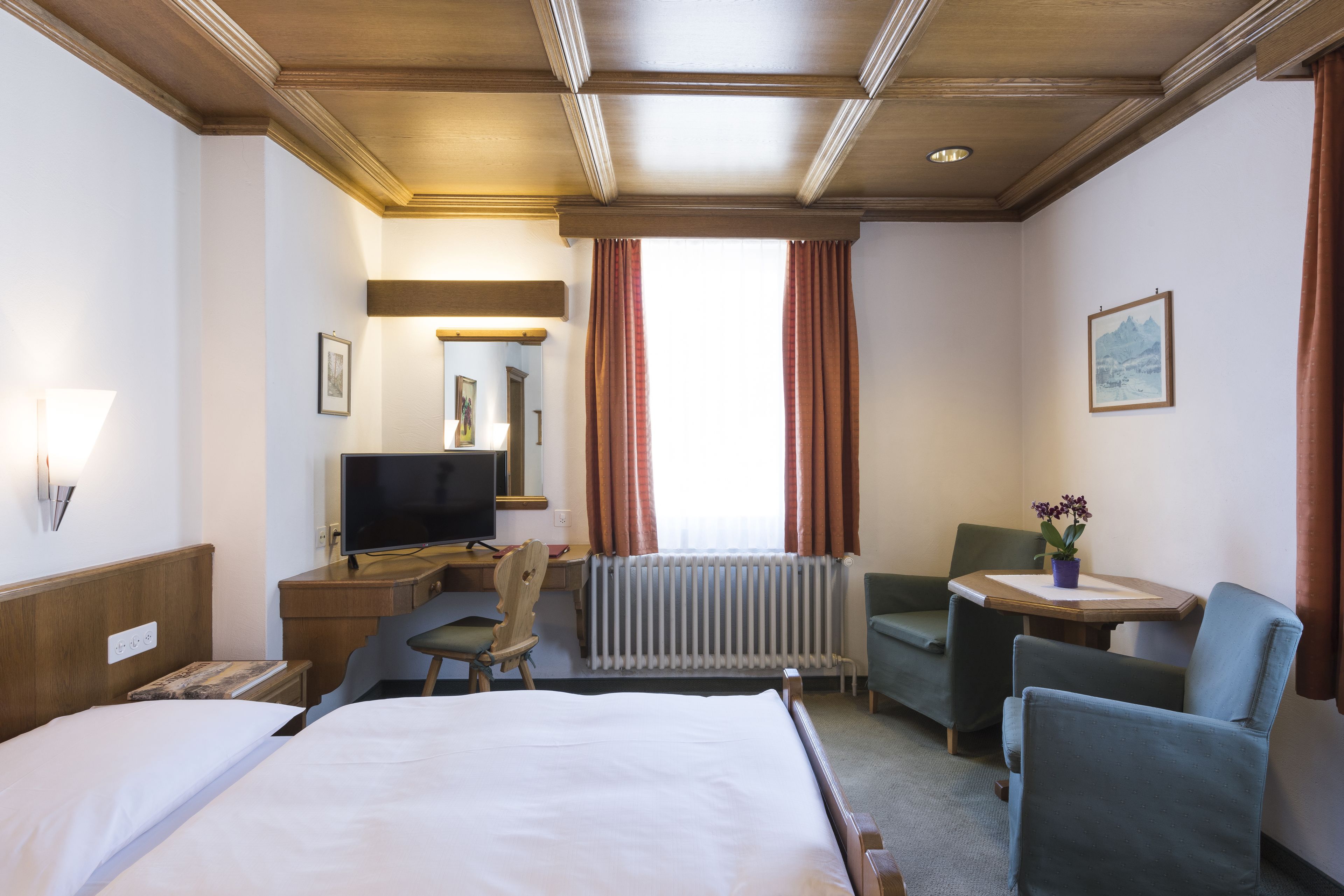A hotel room with a bed, a desk with a computer, a chair, a small table with a plant, and a window with curtains.