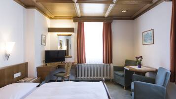 A hotel room with a bed, a desk with a computer, a chair, a small table with a plant, and a window with curtains.