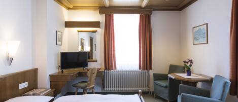 A hotel room with a bed, a desk with a computer, a chair, a small table with a plant, and a window with curtains.