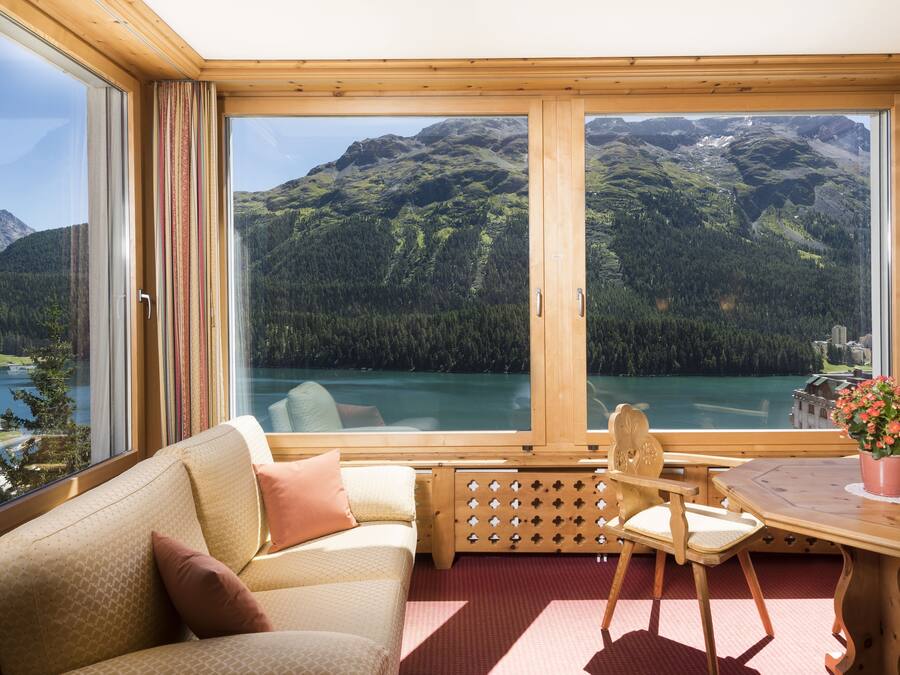 A cozy room with a large window offering a mountain view, a wooden table with a chair, and a sofa with cushions.
