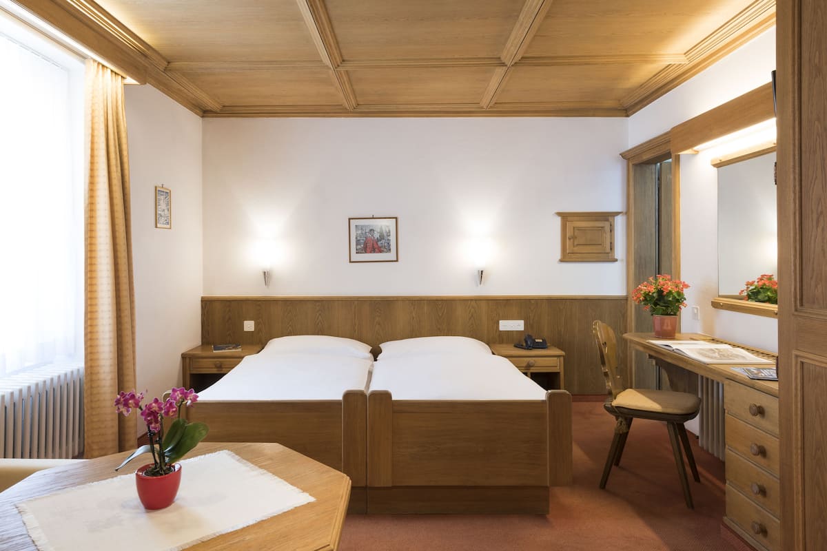A hotel room with two wooden beds, a desk with a chair, a dresser, a mirror, and a window with curtains.