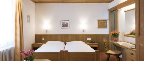 Una habitación de hotel con dos camas de madera, un escritorio con una silla, un aparador, un espejo y un ventanal con cortinas.
