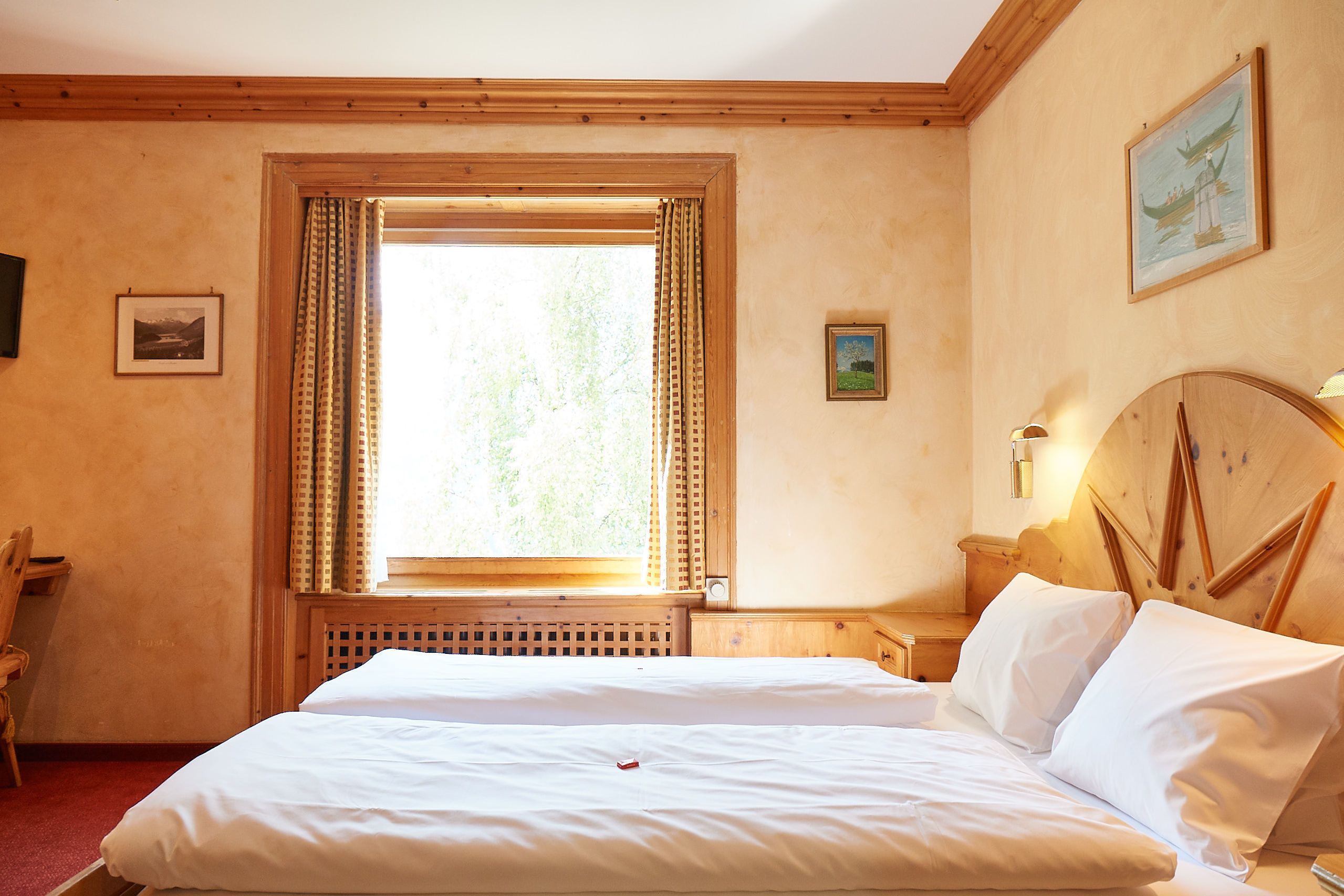 A hotel room with a large bed, a wooden headboard, a window with curtains, a small desk, and a chair.