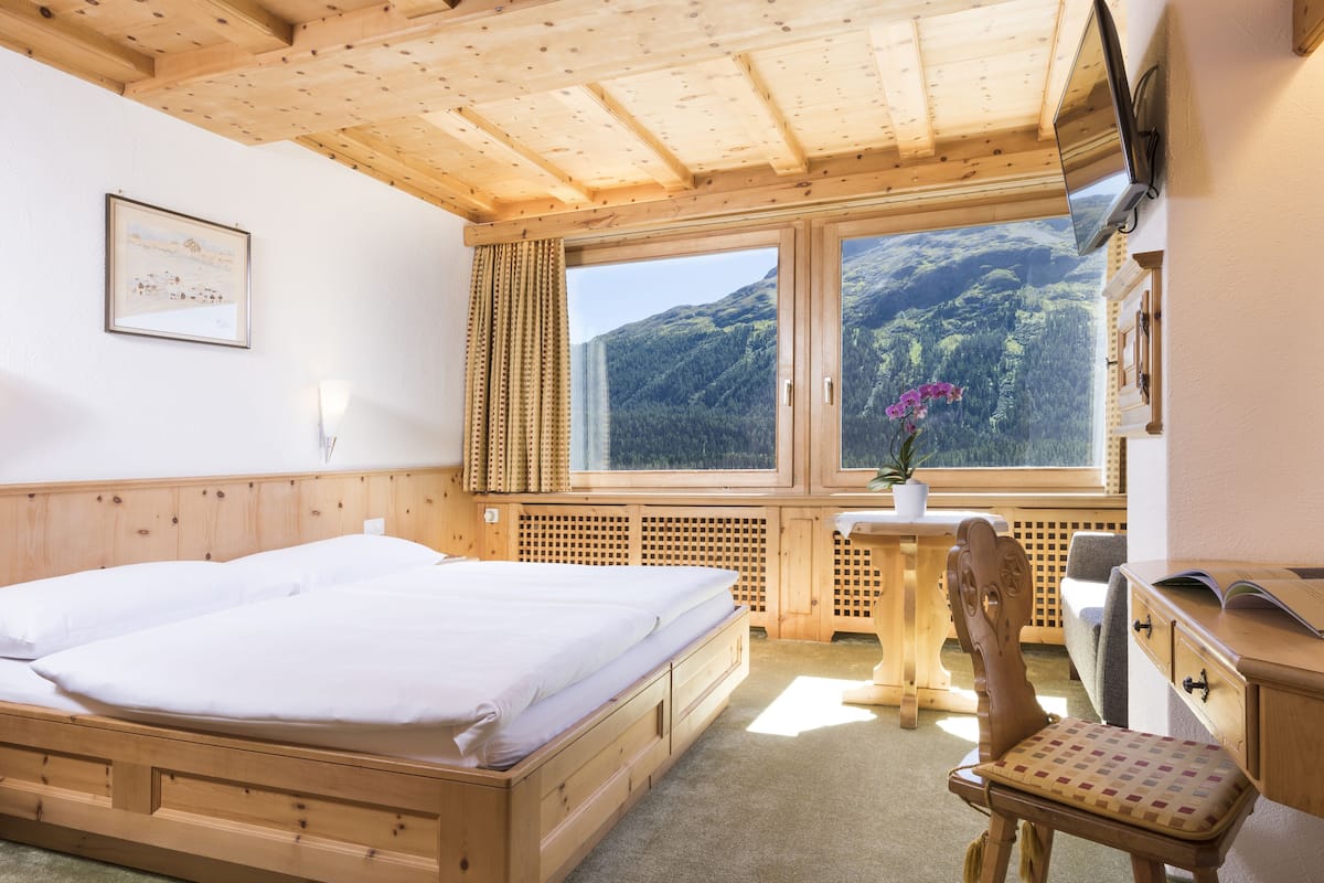 A wooden room with a bed, a desk, a chair, and a window offering a mountain view.
