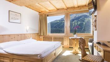 A wooden room with a bed, a desk, a chair, and a window offering a mountain view.