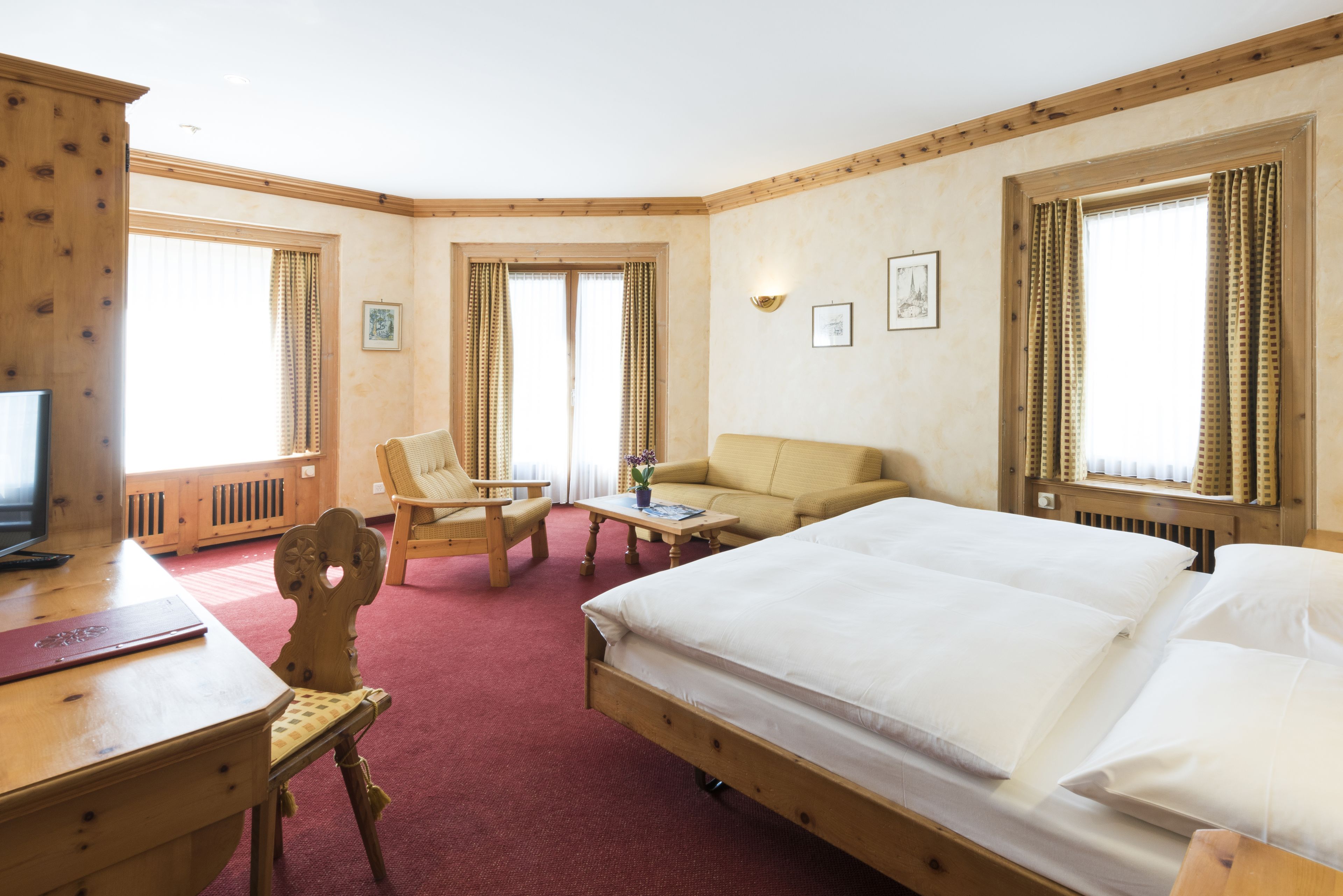 A hotel room with a large bed, a desk with a computer, a chair, a sofa, and two windows with curtains.