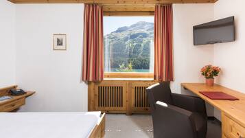 A hotel room with a bed, desk, chair, TV, window with curtains, and a view of mountains.