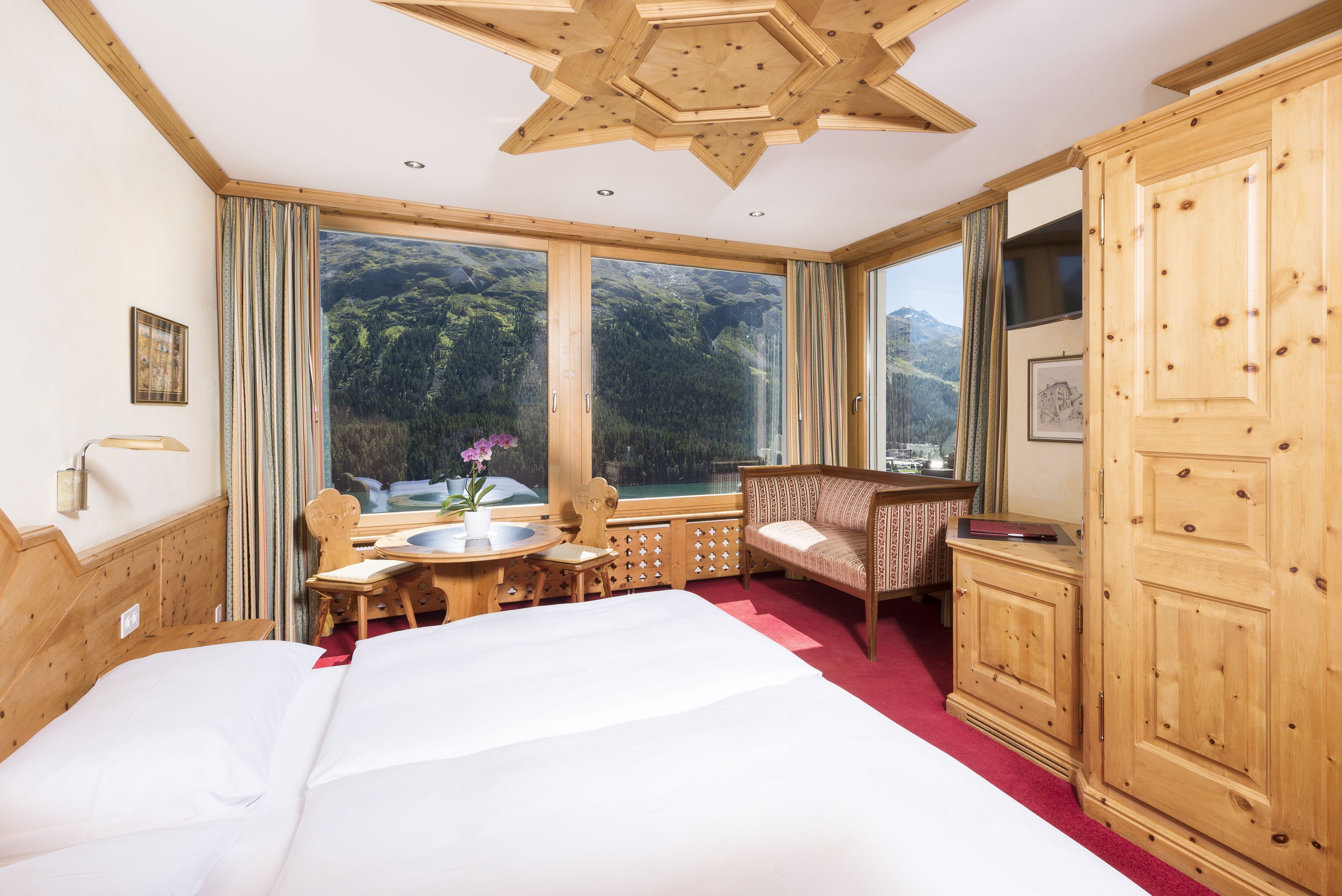 A room with a large bed, wooden furniture, a view of mountains through large windows, and a wooden ceiling.