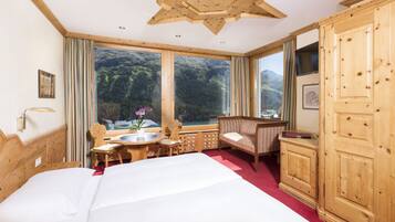 A room with a large bed, wooden furniture, a view of mountains through large windows, and a wooden ceiling.