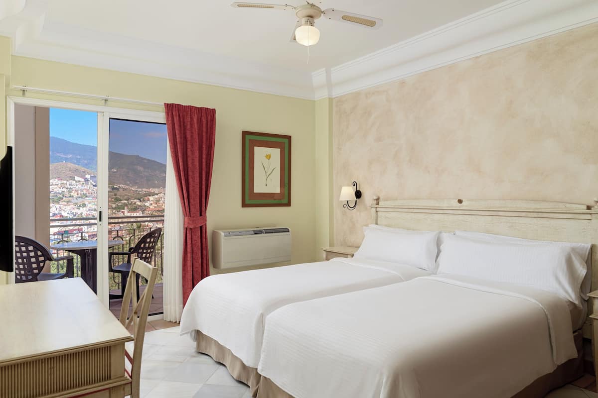A hotel room with a bed, a desk, and a chair. There is a window with a view of a city and mountains.