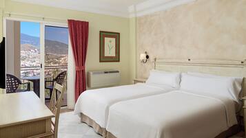 A hotel room with a bed, a desk, and a chair. There is a window with a view of a city and mountains.