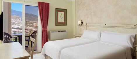 Habitación de hotel con cama, escritorio y silla. Se ve una ventana con vistas a la ciudad y a las montañas.