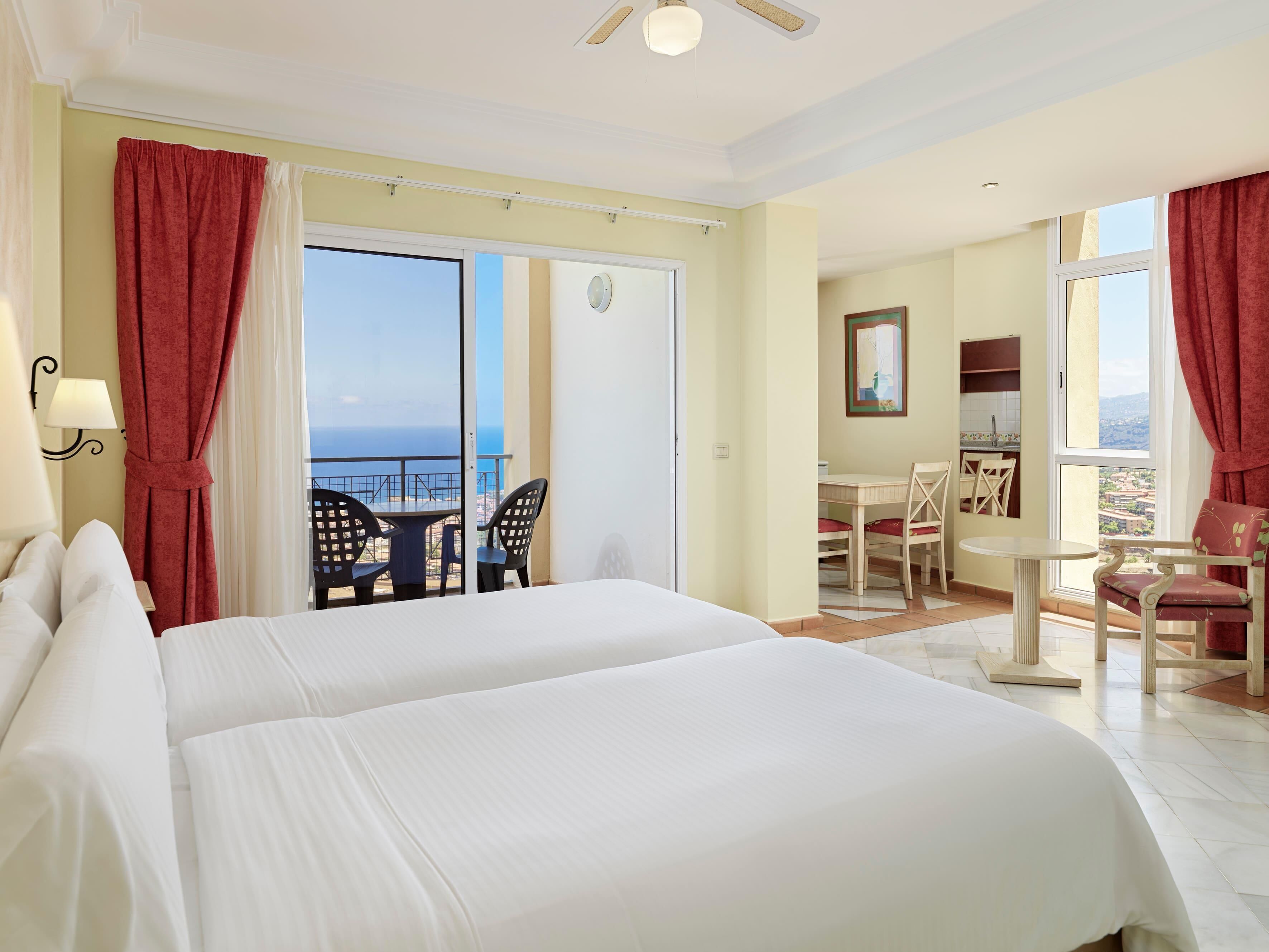 A hotel room with two beds, a balcony with a table and chairs, a kitchenette, and a view of the sea.