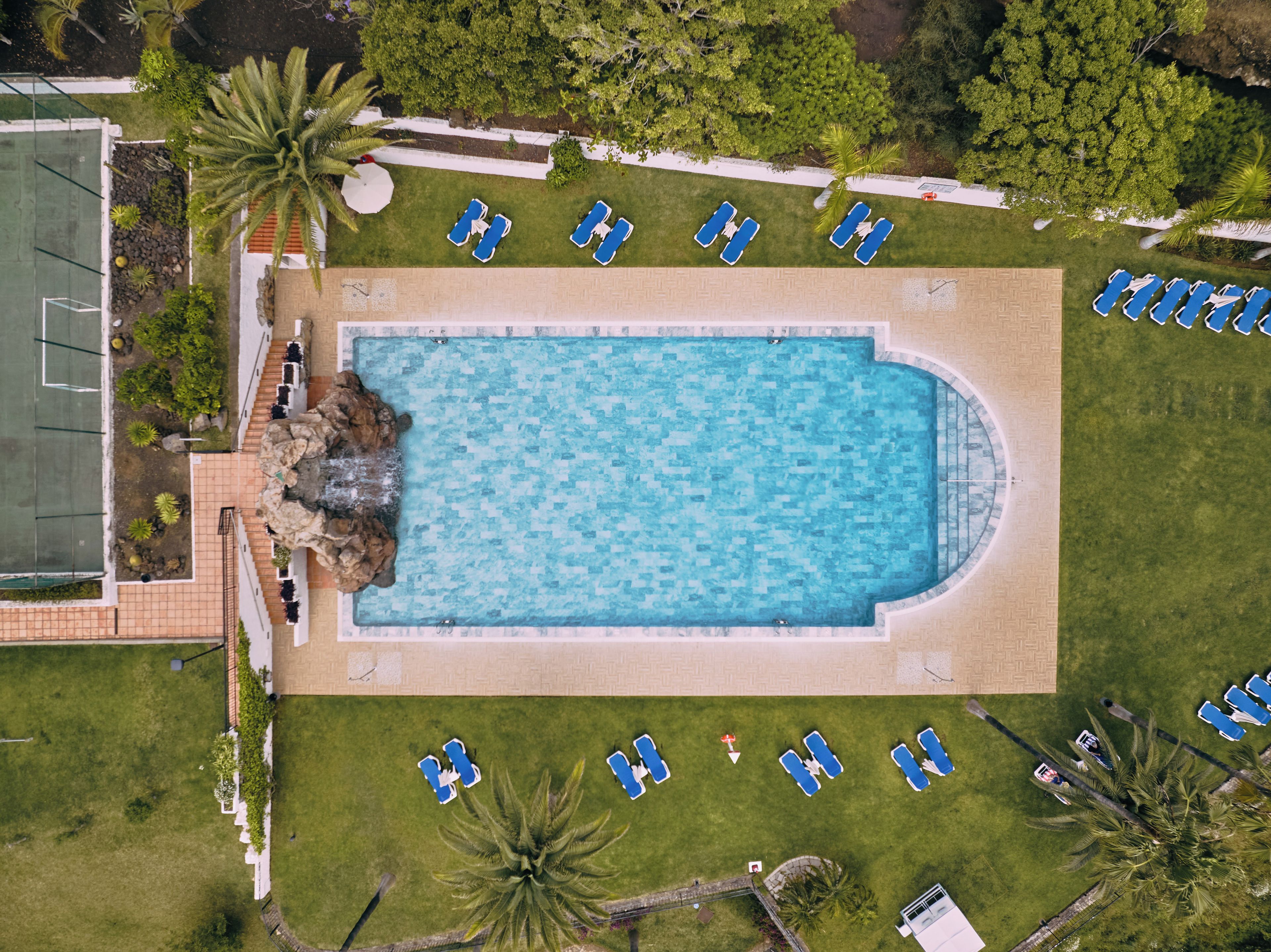 An aerial view of a swimming pool surrounded by lounge chairs, with a tennis court to the left and a paved walkway.