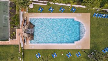 An aerial view of a swimming pool surrounded by lounge chairs, with a tennis court to the left and a paved walkway.
