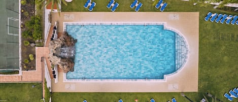 An aerial view of a swimming pool surrounded by lounge chairs, with a tennis court to the left and a paved walkway.
