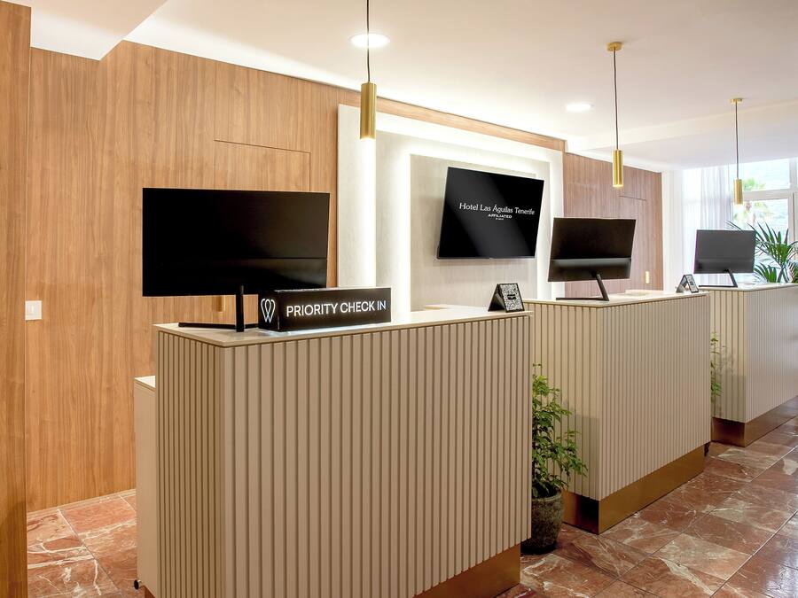 Uma área de recepção de hotel com lambris de madeira, um sinal de check-in prioritário e duas televisões de tela plana na parede.