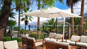 A terrace with wooden furniture and a white umbrella, overlooking a coastal view with palm trees and a distant cityscape. - Hotel Las Aguilas Tenerife, Affiliated by Melia (Puerto de la Cruz)