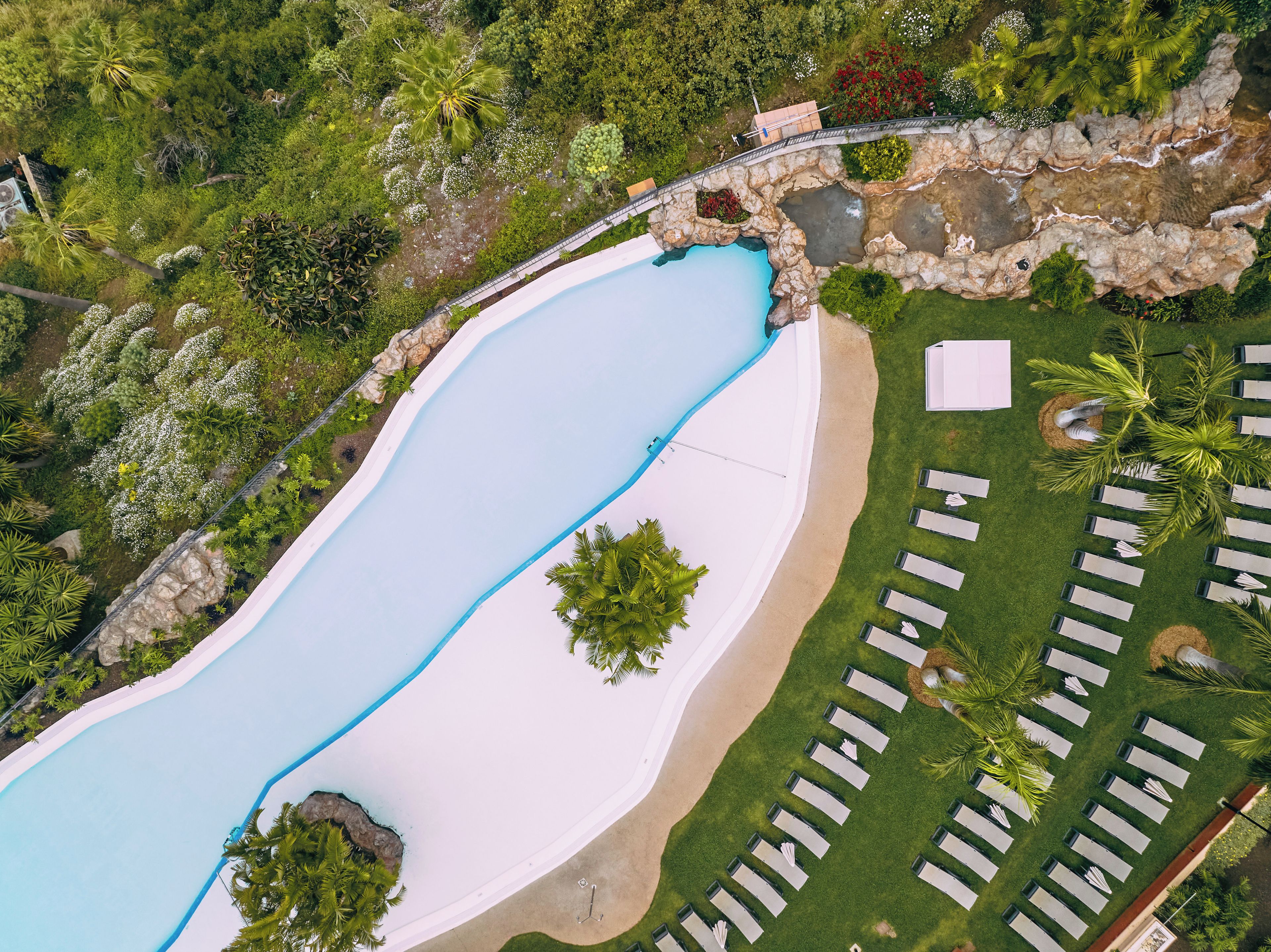 An aerial view of a resort with a large swimming pool, a bridge, and a row of sunbeds.