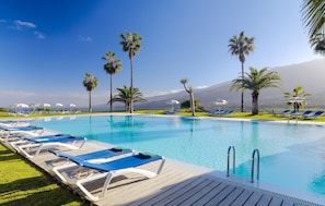 Zona de piscina con sillas de descanso y palmeras, rodeada por una valla y una montaña al fondo.