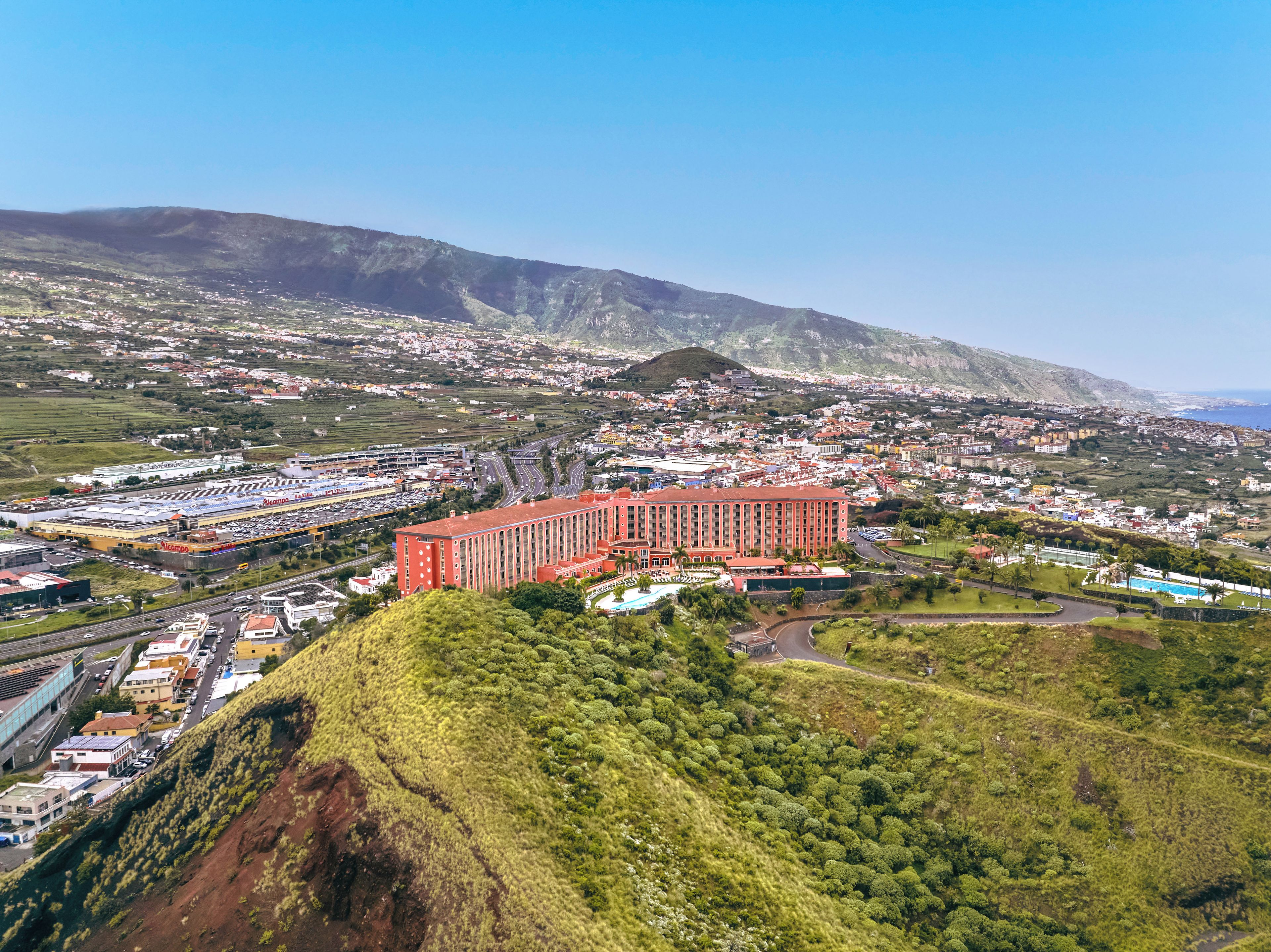 A large red building with multiple floors, surrounded by a hilly landscape and a coastal view.